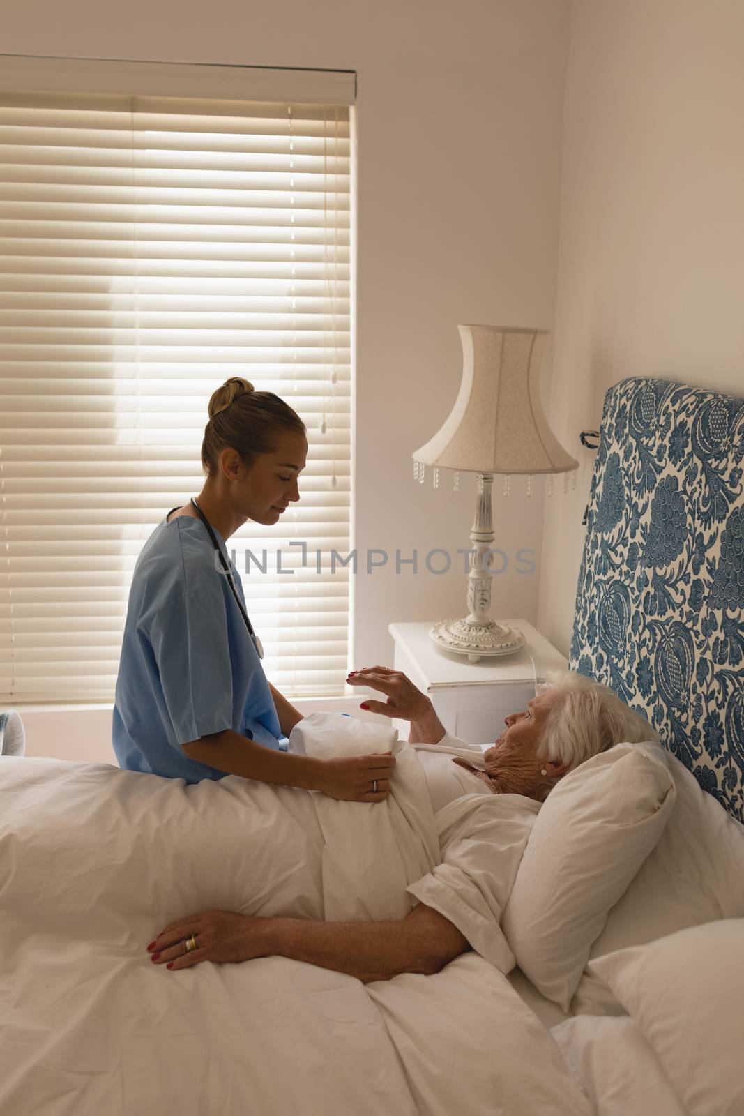 Side view of a senior woman lying while interacting with beautiful female doctor in bedroom at home