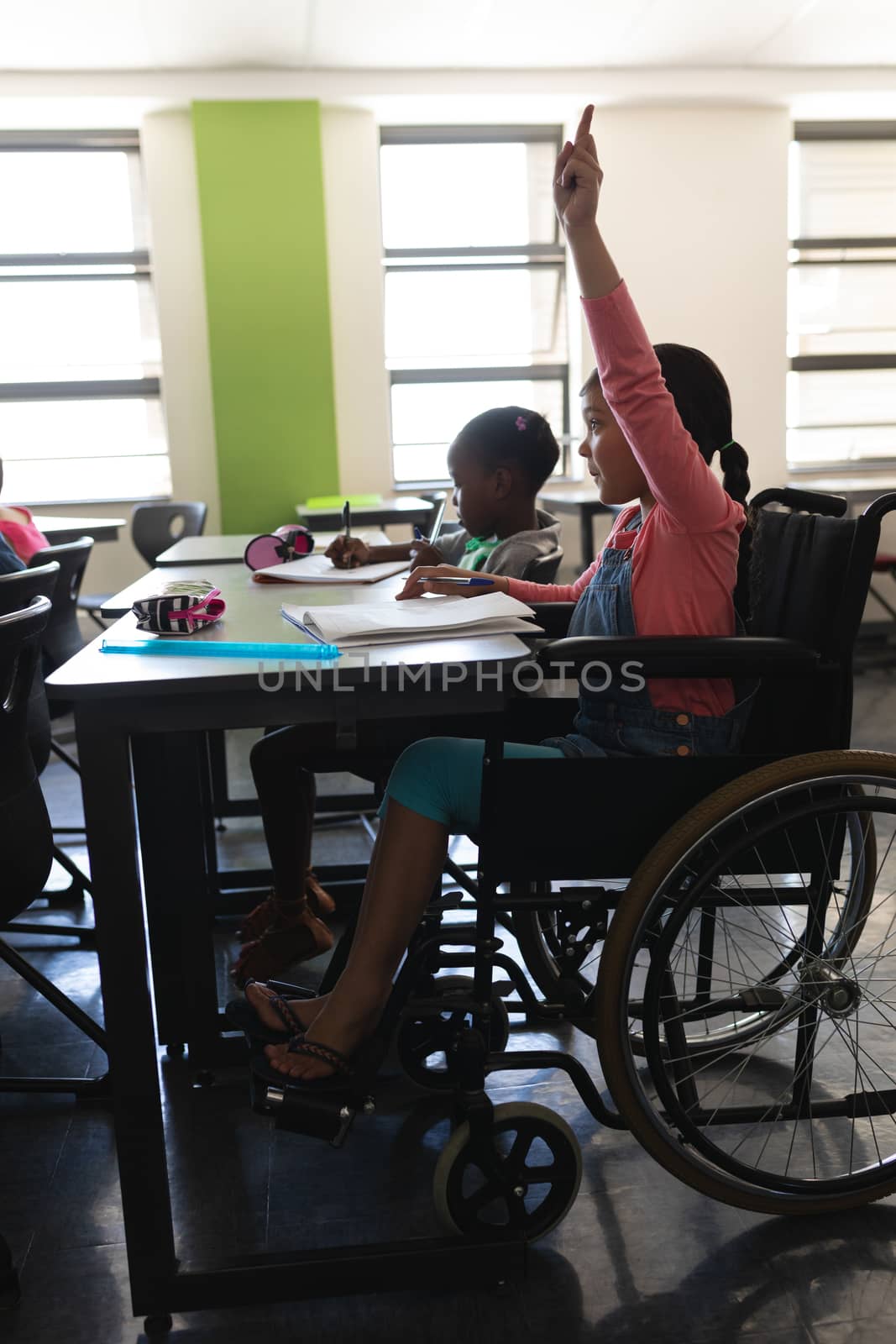Side view of disable schoolgirl raising hand and sitting at desk in classroom  by Wavebreakmedia