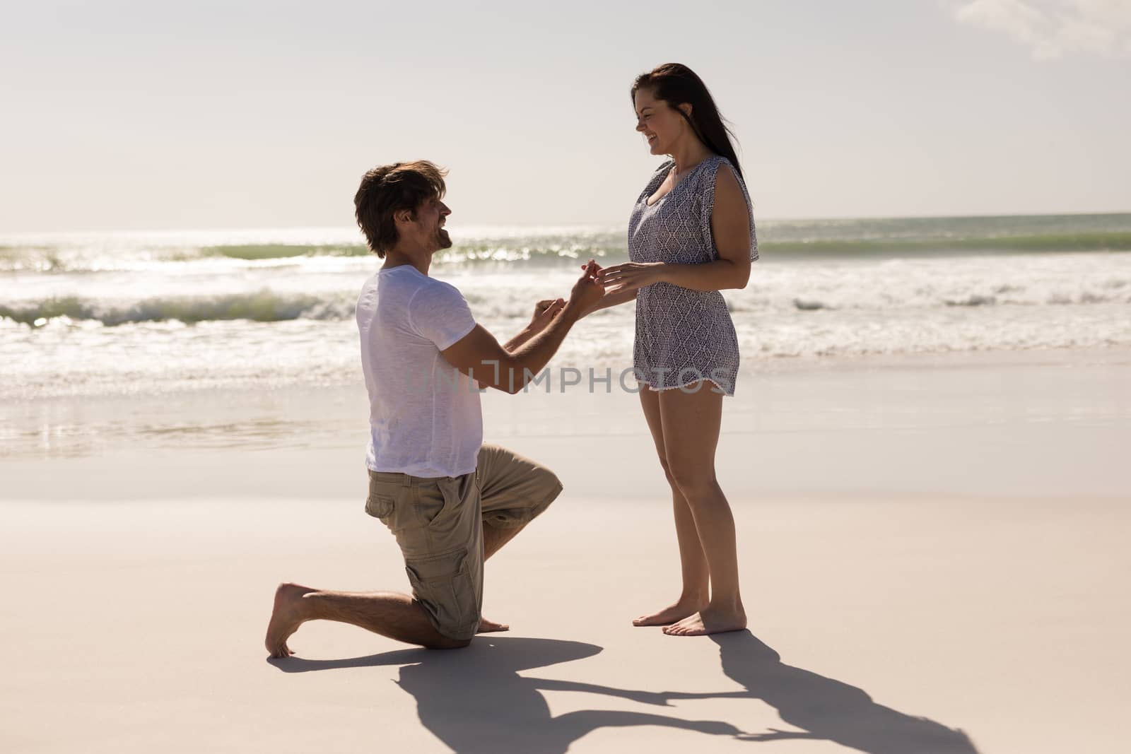 Side view of romantic young man proposing to a woman on his knee at beach in the sunshine