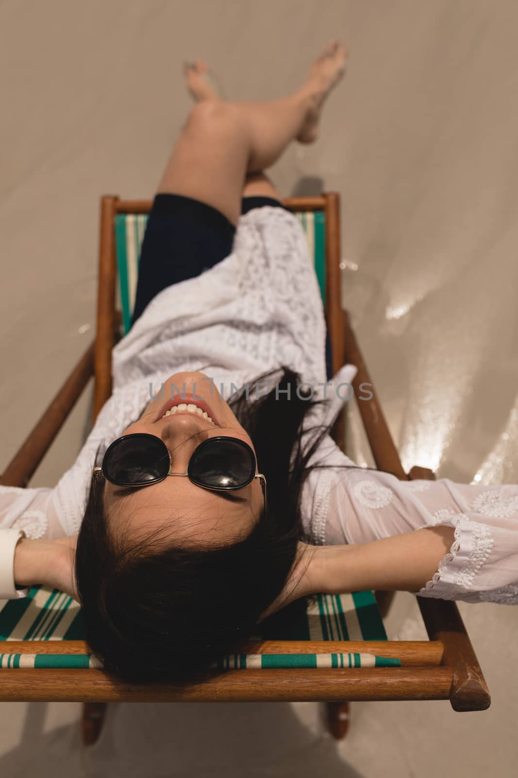 Overhead of beautiful young woman with hand behind head sitting on sun lounger and looking at camera on beach