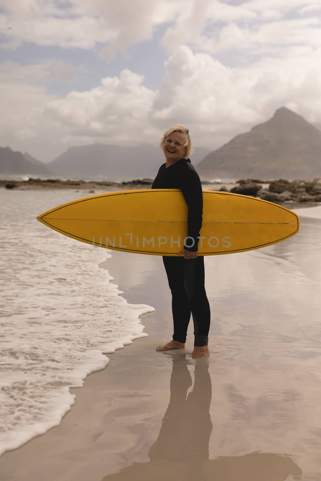 Side view of an active senior woman standing with surfboard on the beach