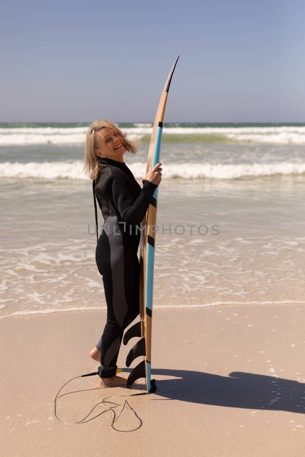 Side view of active senior female surfer standing with surfboard on the beach