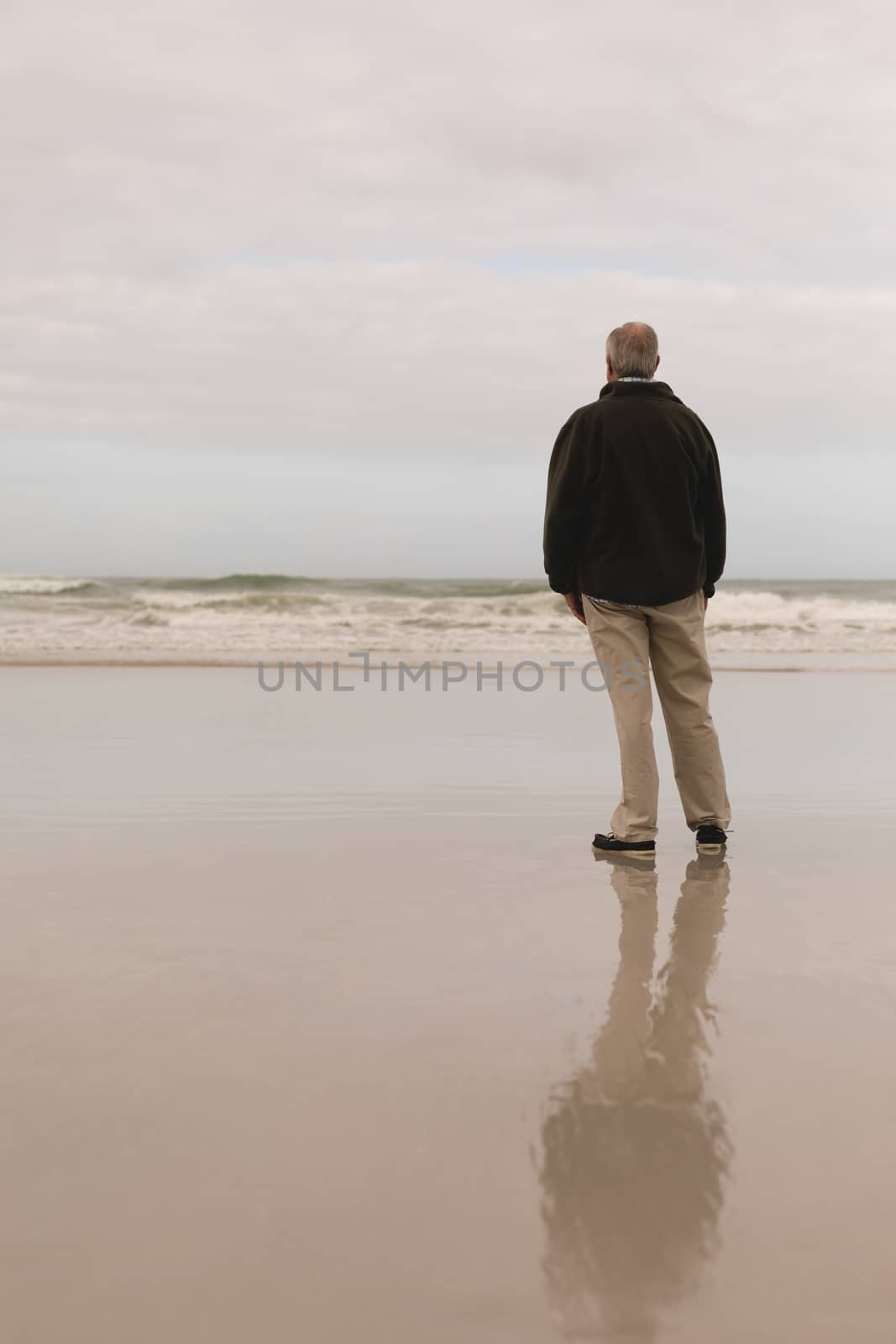 Back view of active senior man standing front of ocean the hands in his pocket. He seems relaxed