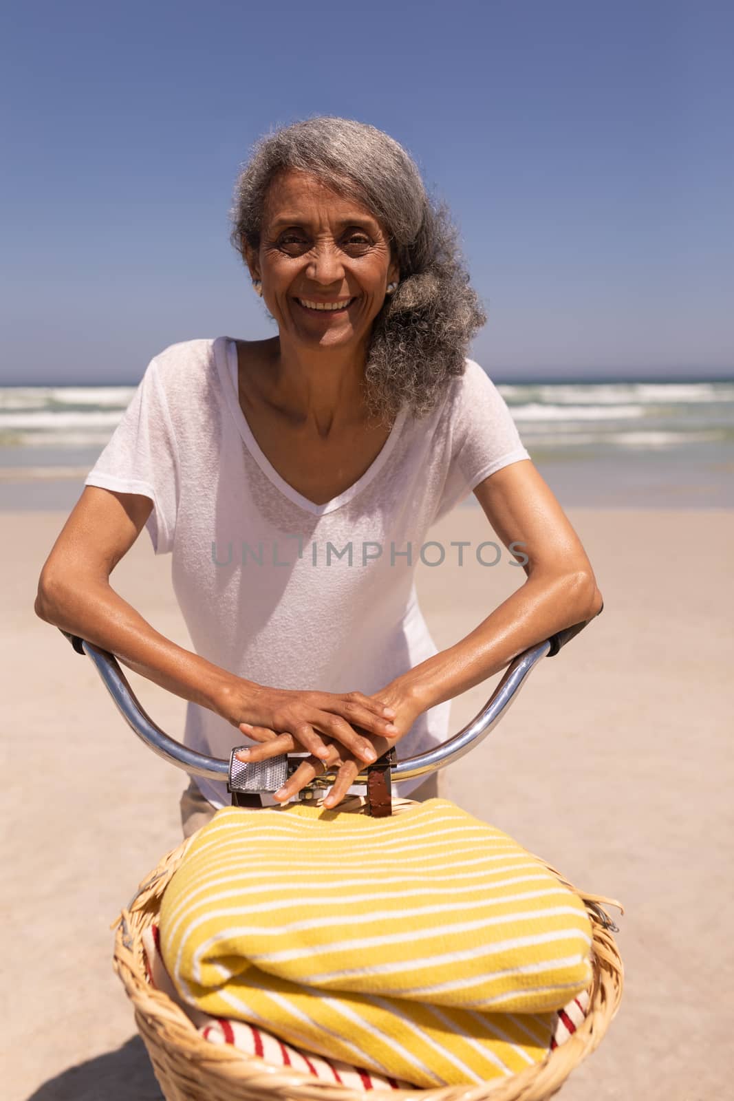 Front view of happy senior woman with bicycle standing on beach in the sunshine