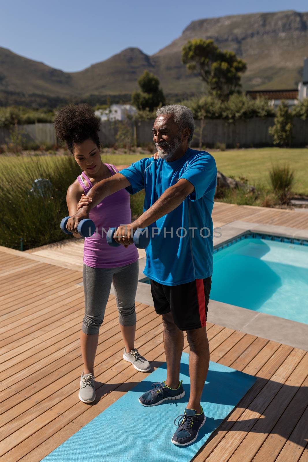 Front view of a female  African American trainer assisting senior man in performing exercise next to the swimming pool in the backyard of home