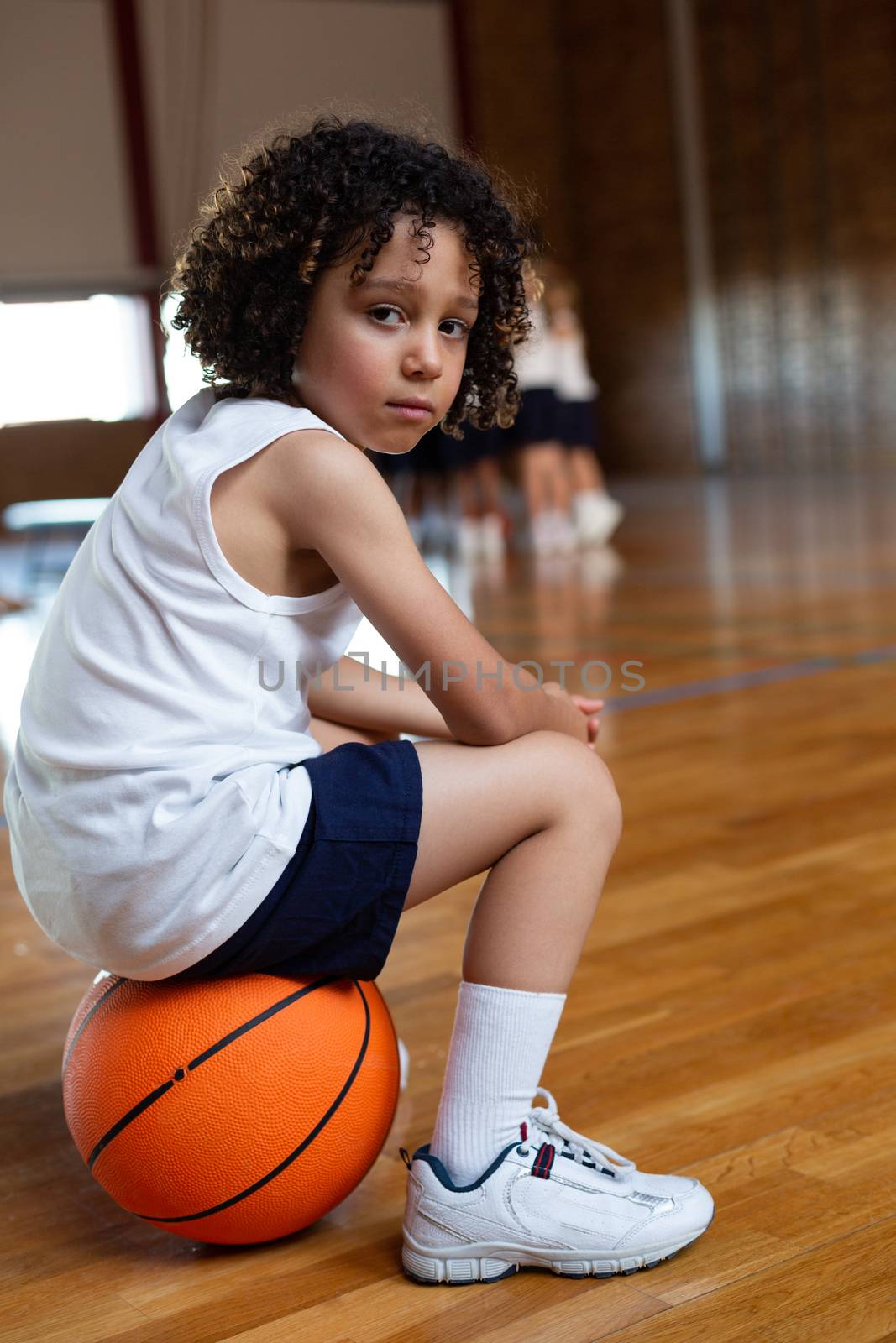 Side view of a mixed-race schoolboy sitting on a basketball and looking at camera in basketball court at school