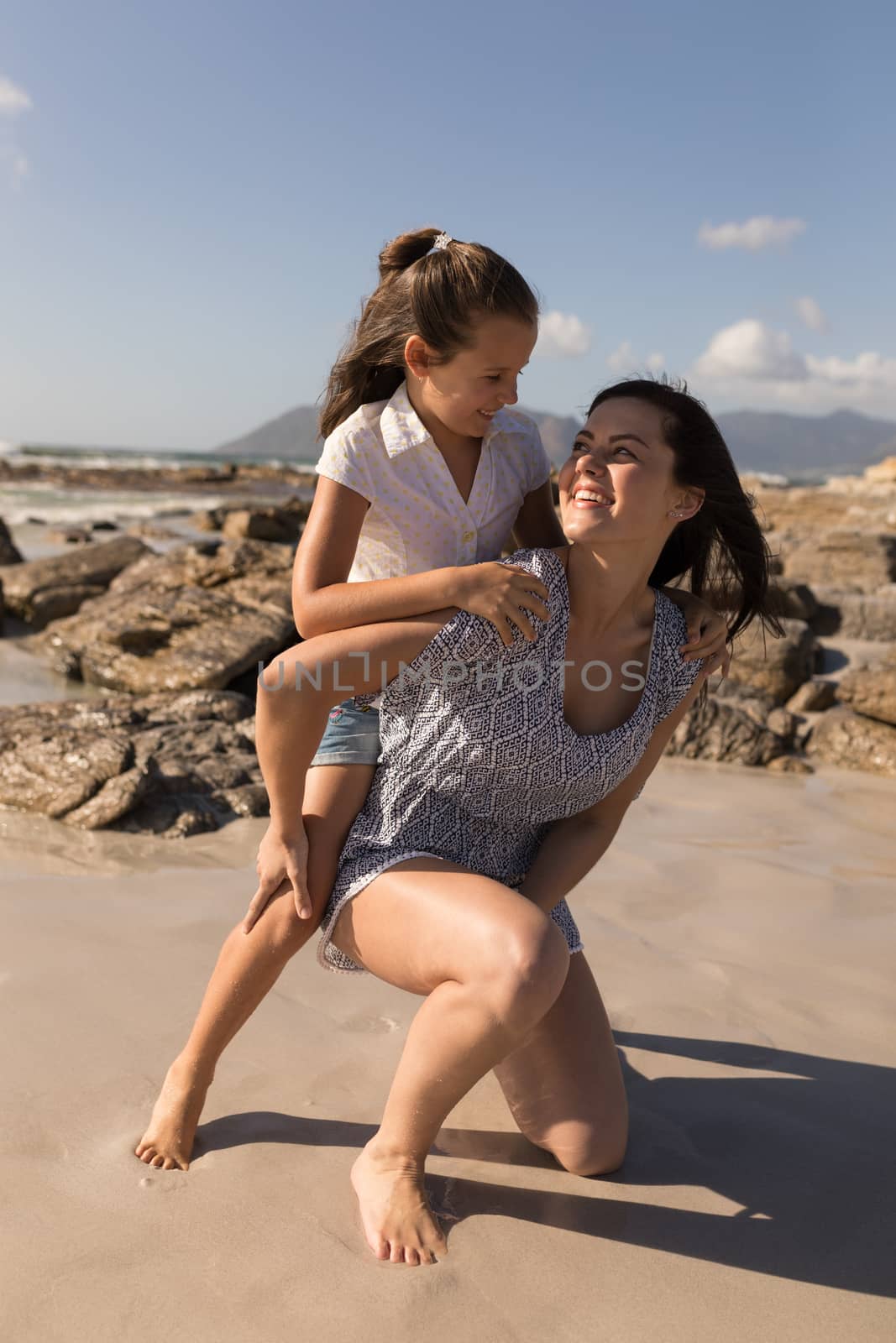Front view of beautiful young mother giving piggyback to her daughter on beach