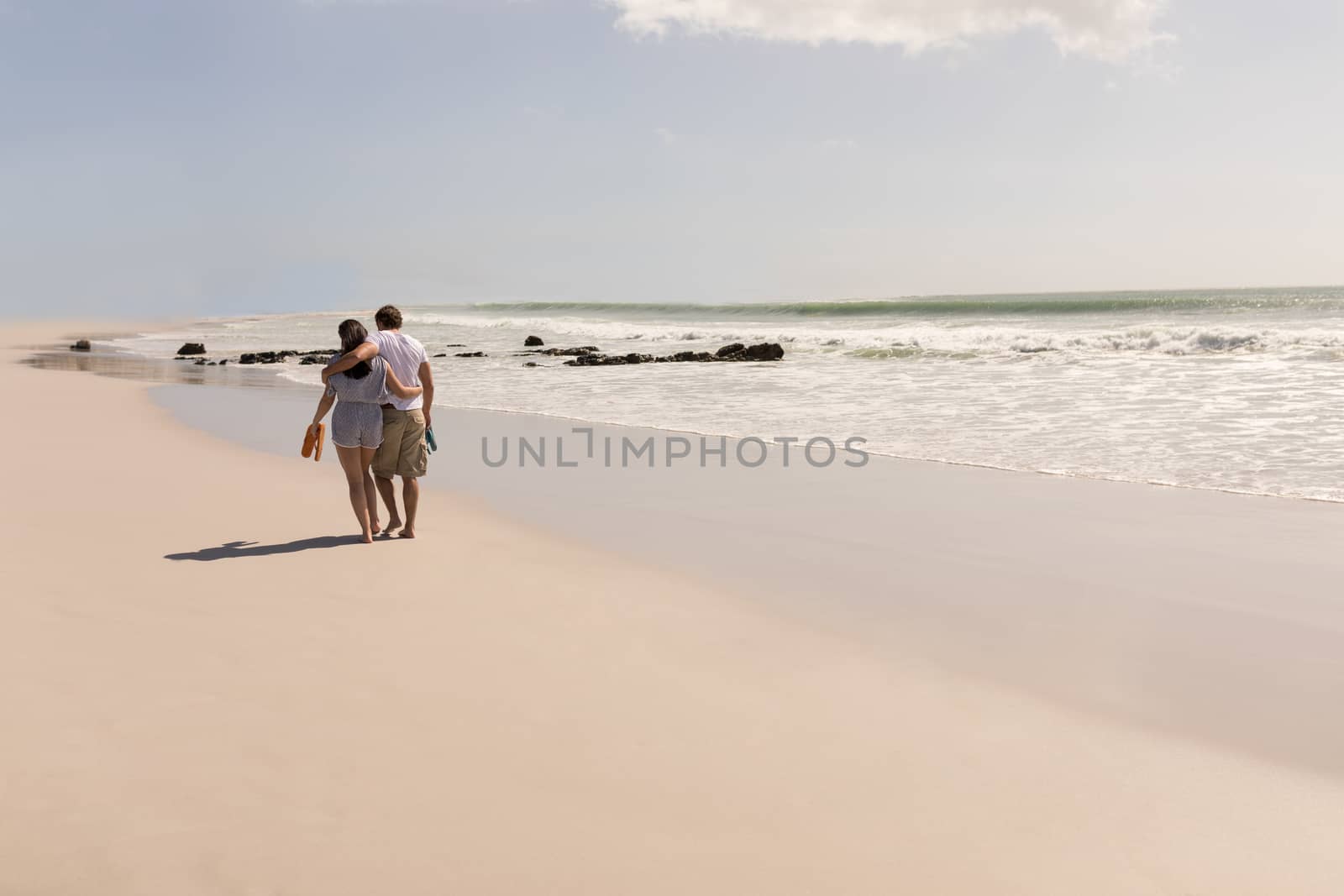 Rear view of romantic young couple walking with beach footwear on beach in the sunshine
