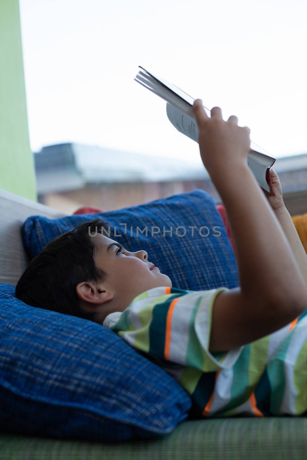 Side view of a Caucasian schoolboy lying on sofa and reading a book in library at elementary school