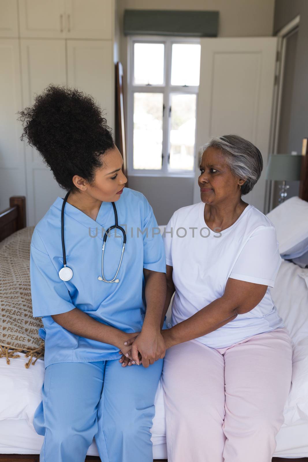 Front view of young pretty African American female doctor comforting senior African American woman in bedroom at home