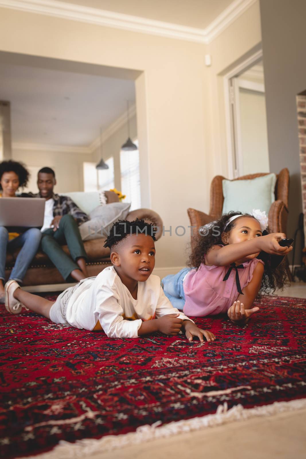 Side view of happy African American sibling lying on floor and watching television while parents using laptop on sofa in a comfortable home