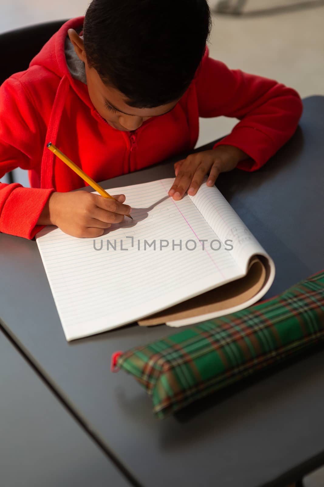 High angle view of cute mixed-race schoolboy writing in a  notebook at desk in classroom at elementary school