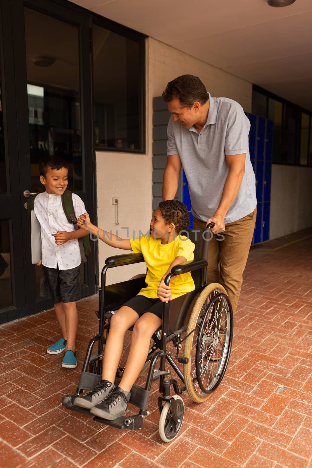 Front view of a cute mixed-race disabled schoolboy in wheelchair with handsome Caucasian male teacher and cute mixed-race classmate in the corridor at school