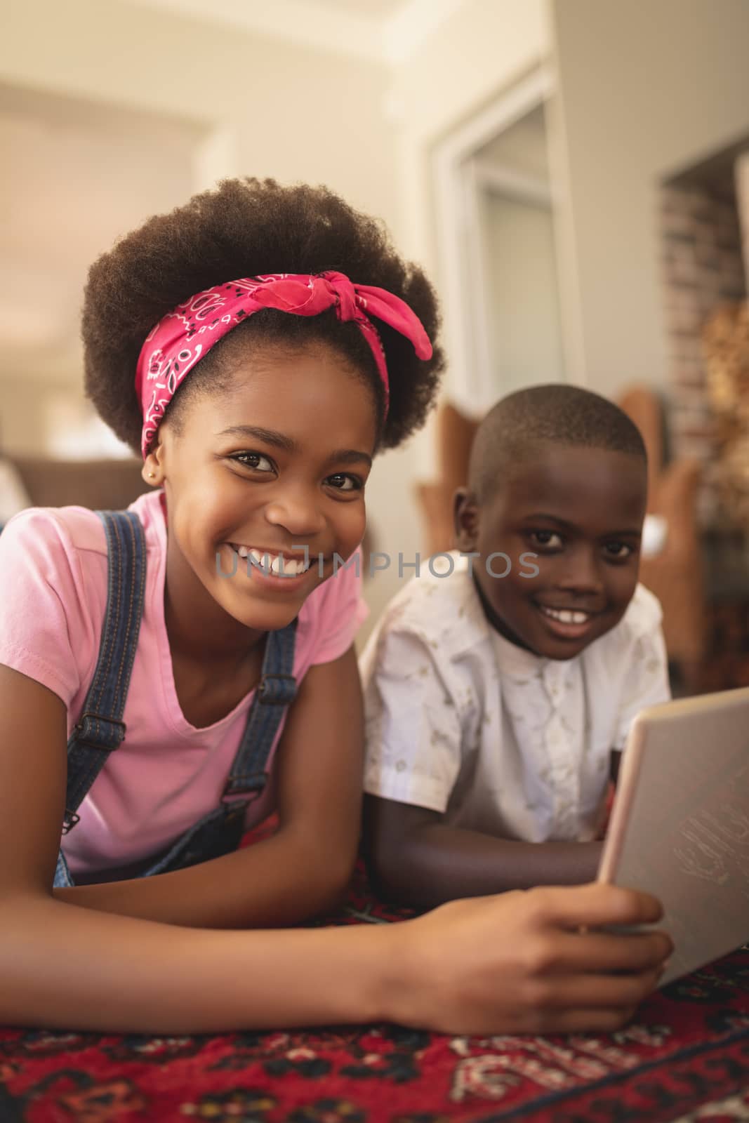 Happy African American sibling lying on floor with digital tablet and looking at camera in a comfortable home