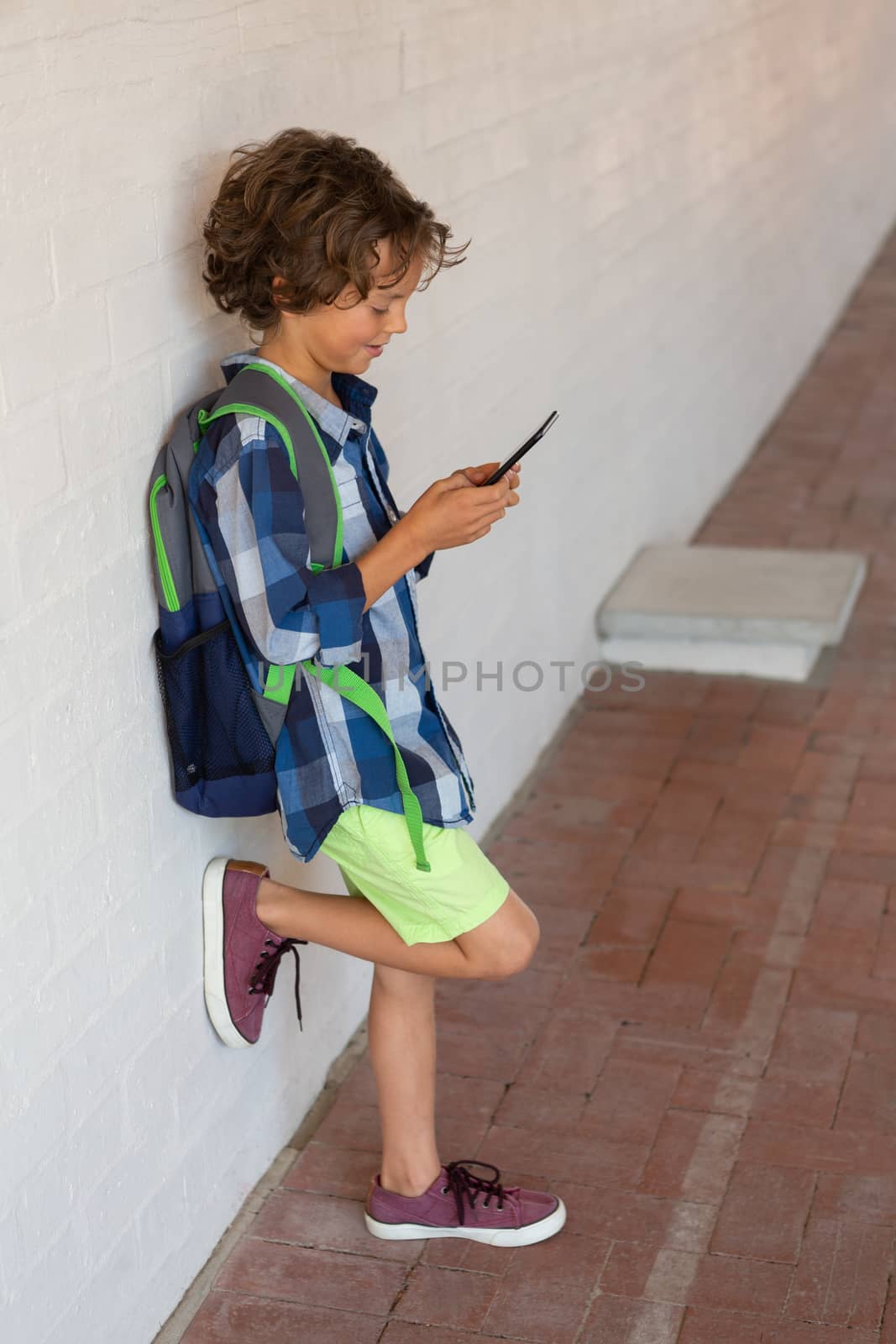 Side view of a Caucasian schoolboy using his mobile phone while leaning against a wall in the corridor at school