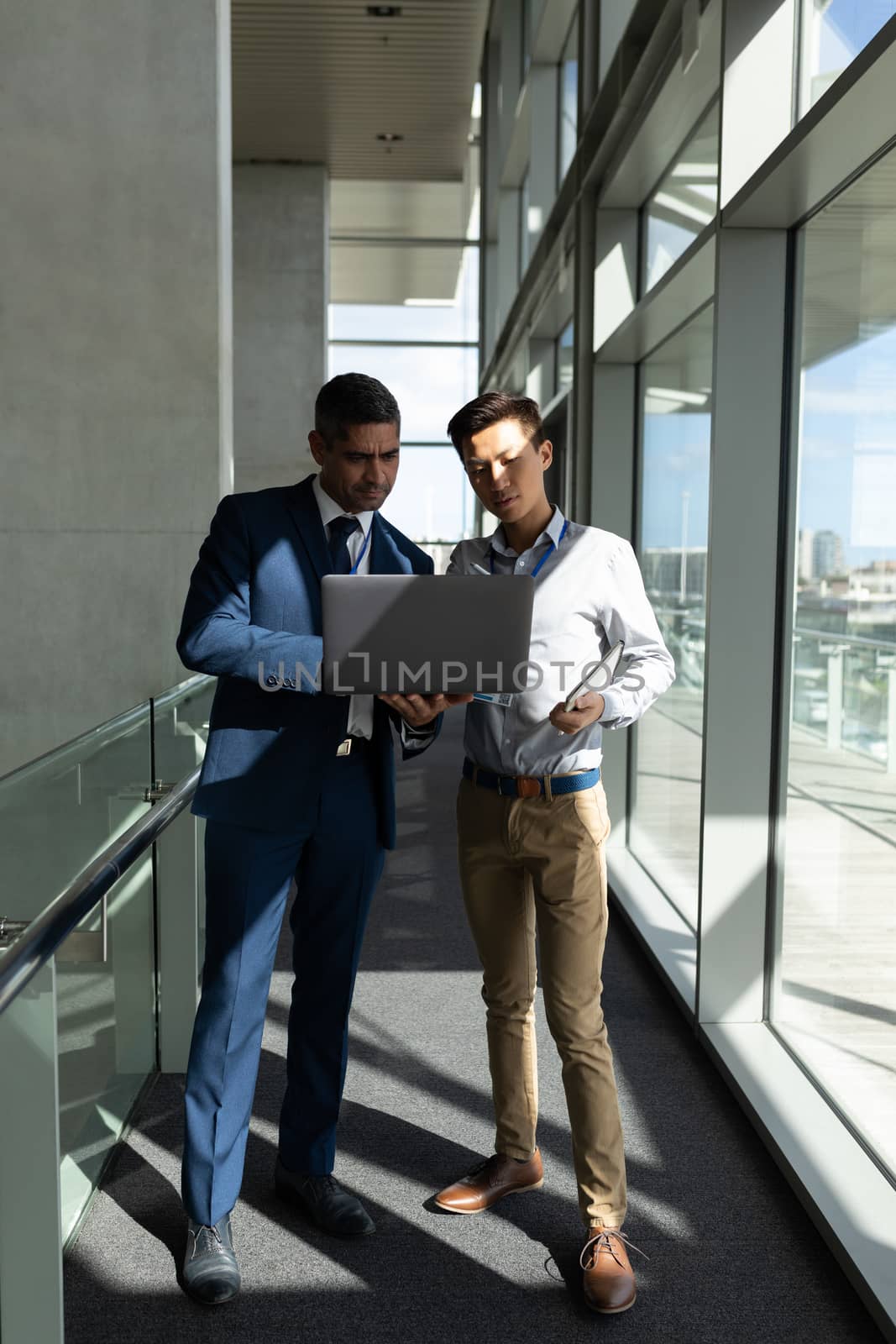 Front view of two businessman standing on the first floor walkway and working on a laptop in office against view on street in background