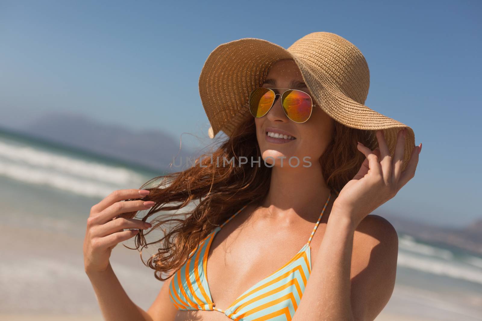 Front view of happy pretty young Caucasian woman in bikini with hat and sunglasses standing on the beach. She seems happy