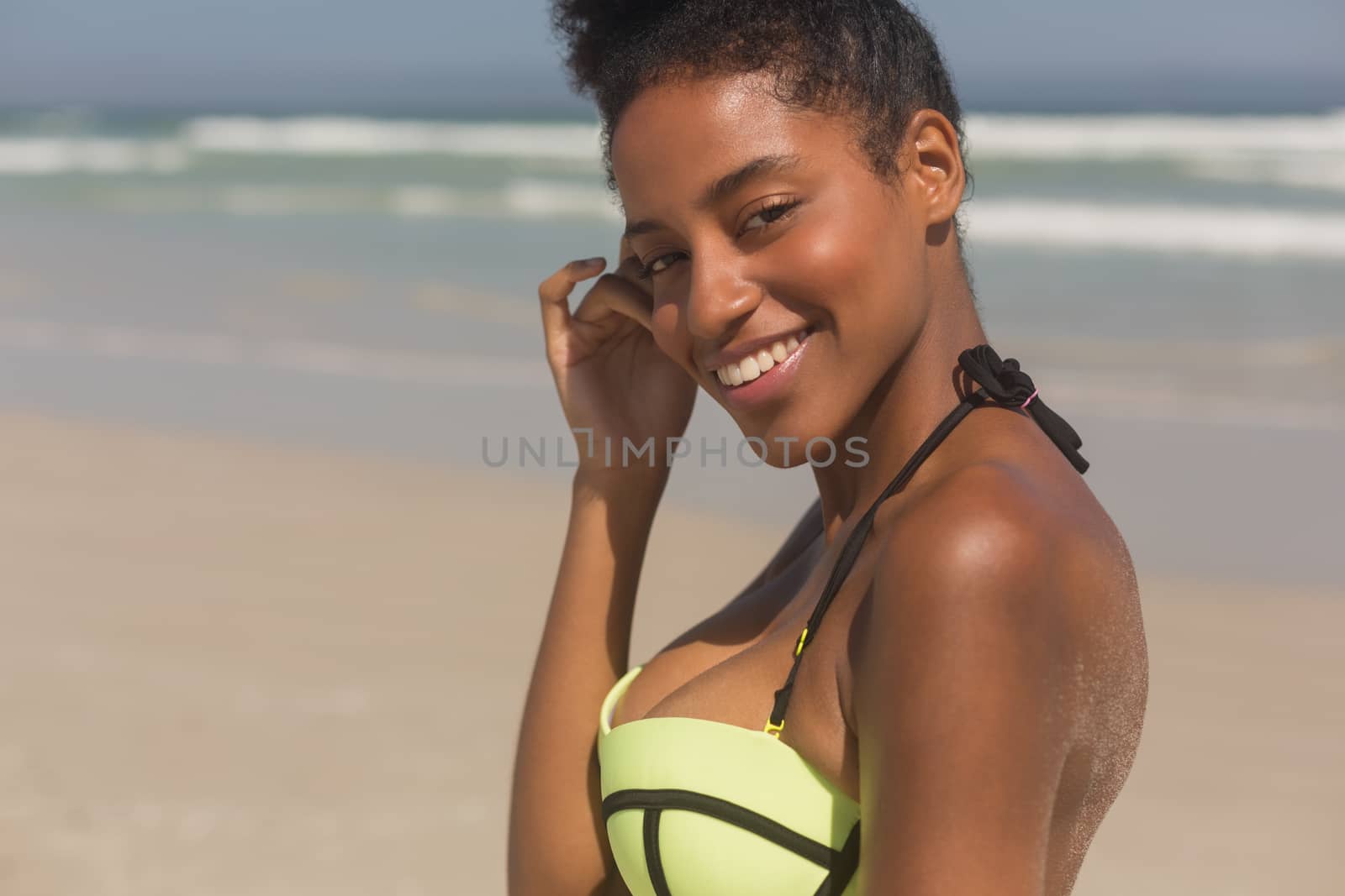Portrait of happy young African American woman in green bikini looking at camera on the beach. She is smiling and looking at camera