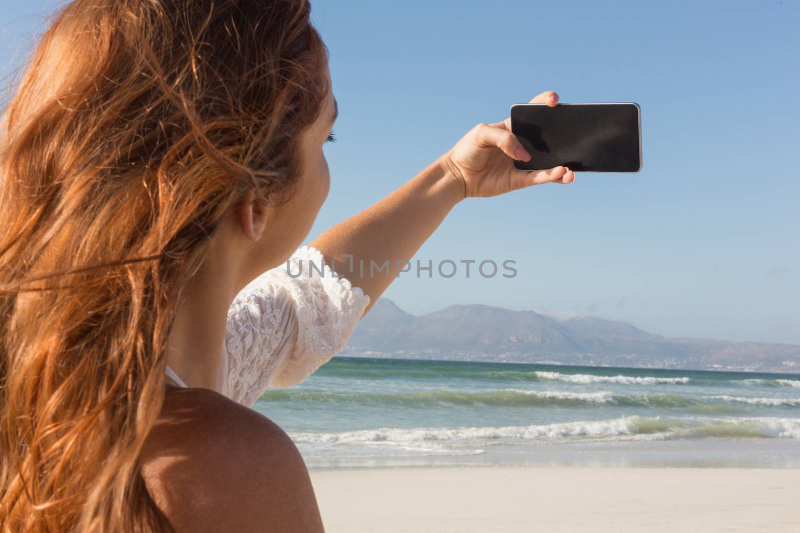 Rear view of beautiful young Caucasian woman taking selfie with her mobile phone standing on the beach. she seems happy
