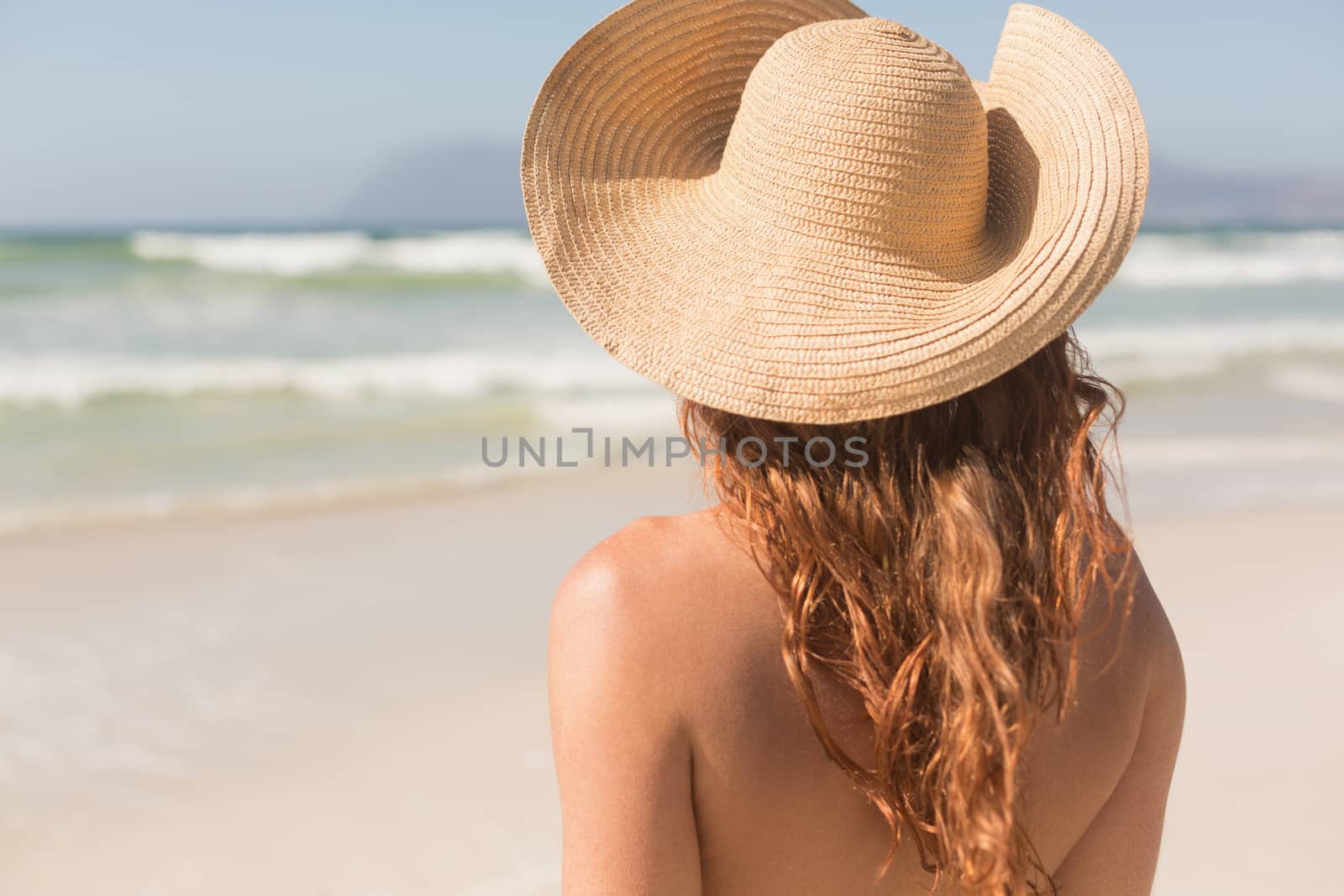 Rear view of young Caucasian woman with hat standing on the beach. She is watching the ocean