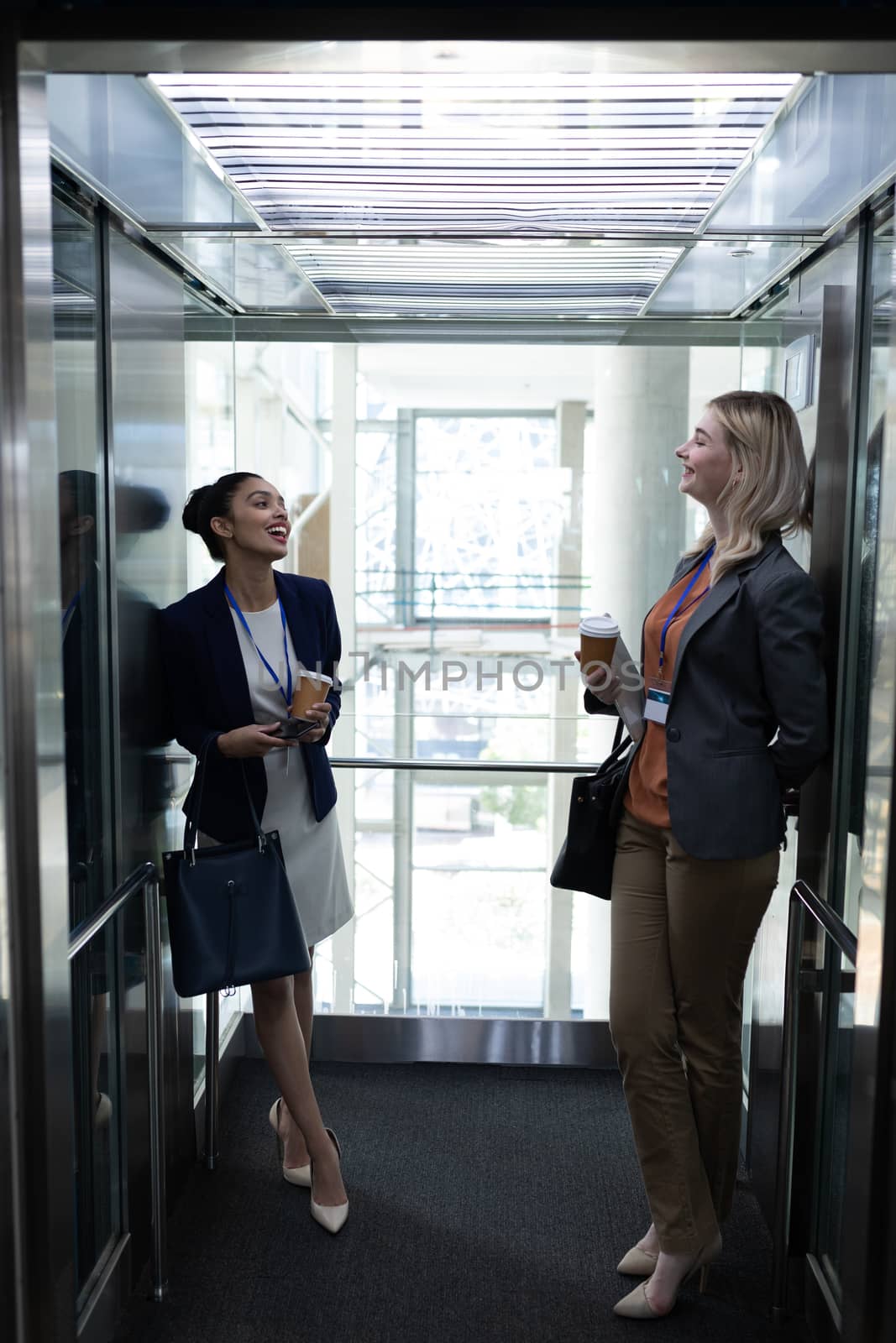 Multi-ethnic businesswomen with coffee cup interacting with each other in office elevator by Wavebreakmedia