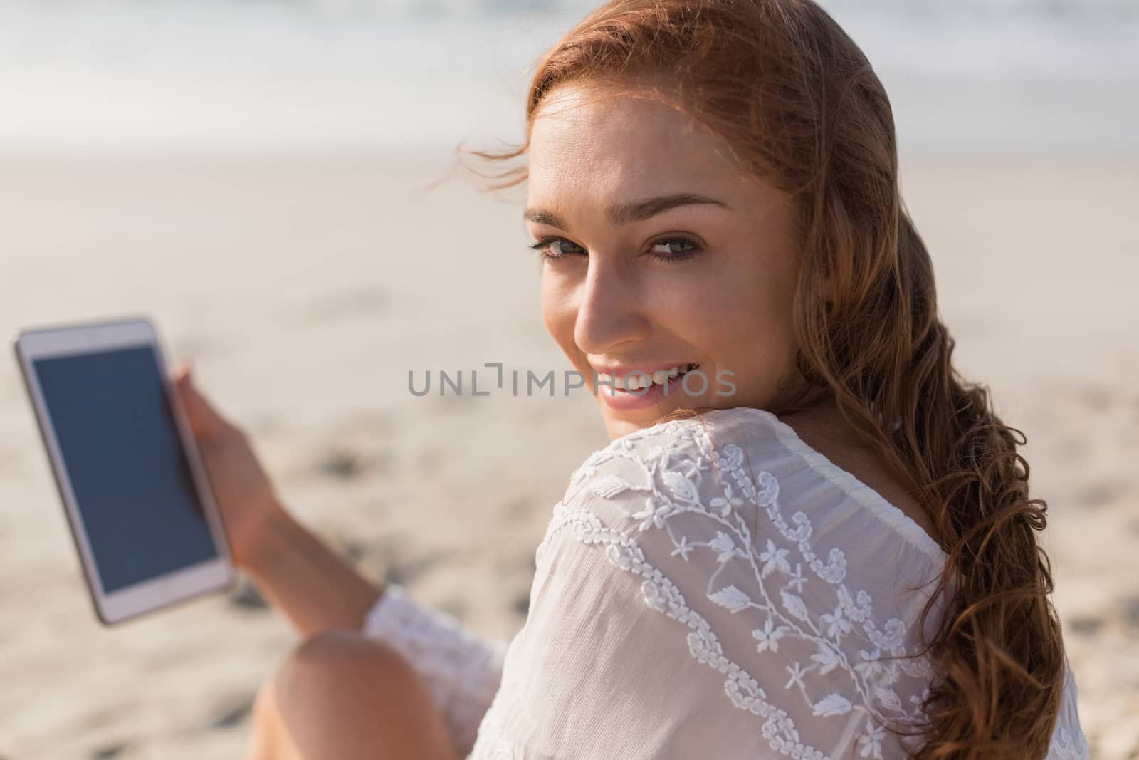 Portrait of beautiful happy young Caucasian woman using digital tablet sitting on the beach. She is looking and smiling at camera