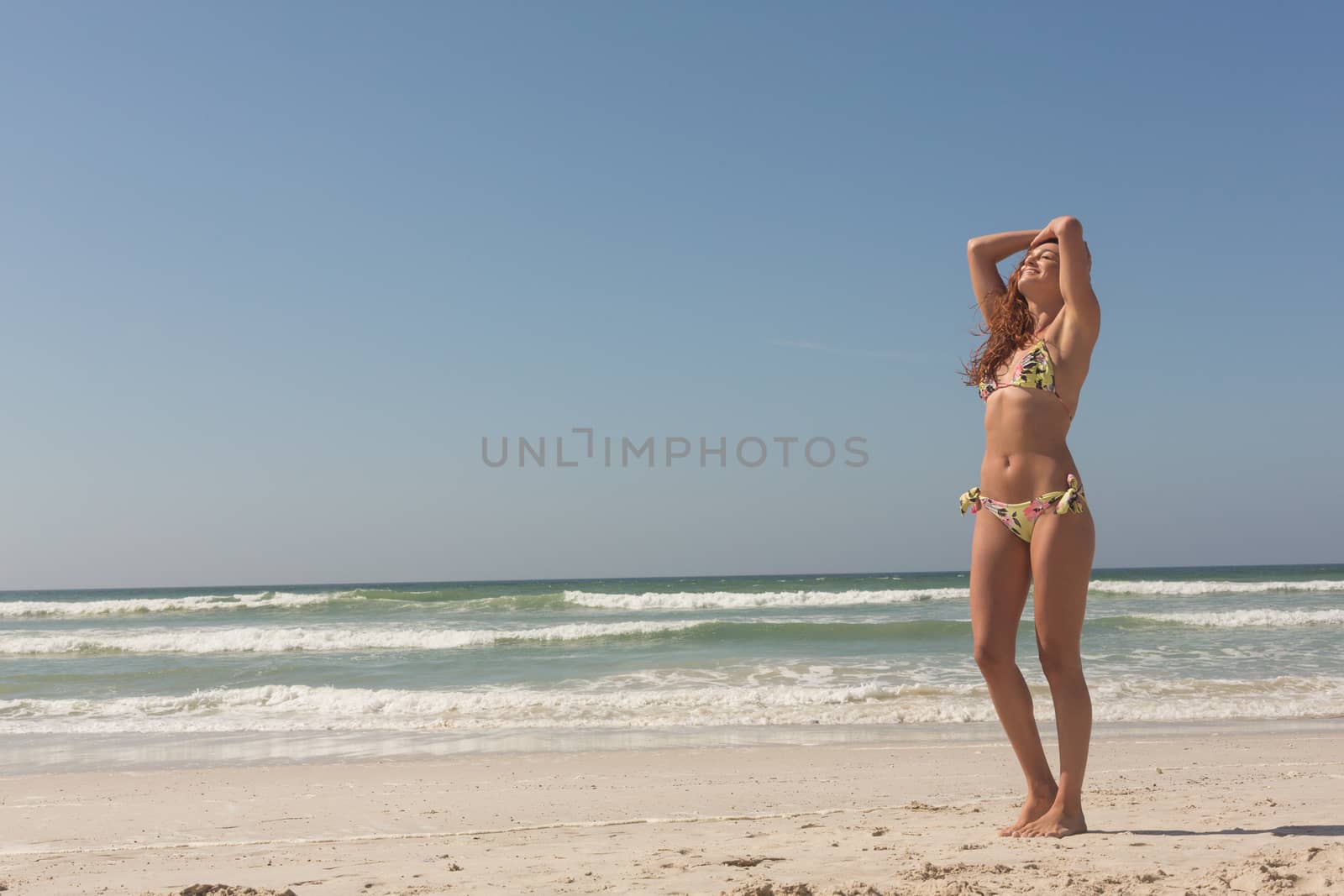 Front view of beautiful young Caucasian woman in bikini standing on the beach. She seems relaxed with her arms above the head