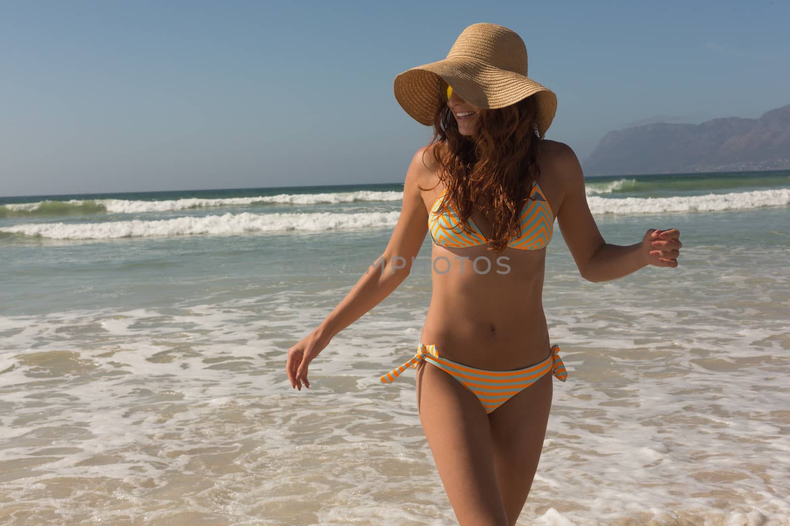 Front view of beautiful young Caucasian woman in bikini and hat walking on the beach. She is smiling