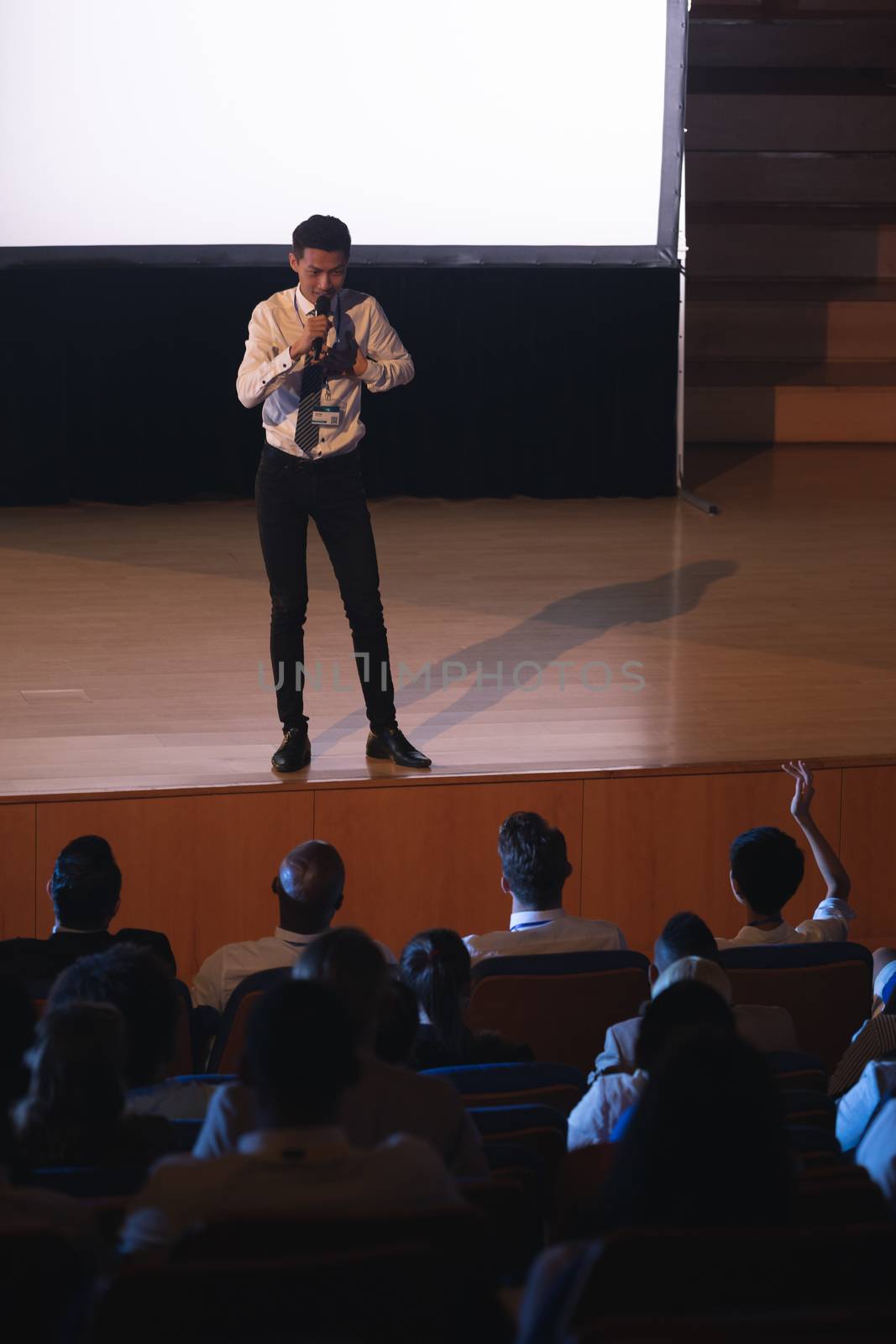 High view of young Asian businessman standing and giving presentation in auditorium while audience raising hand for asking query