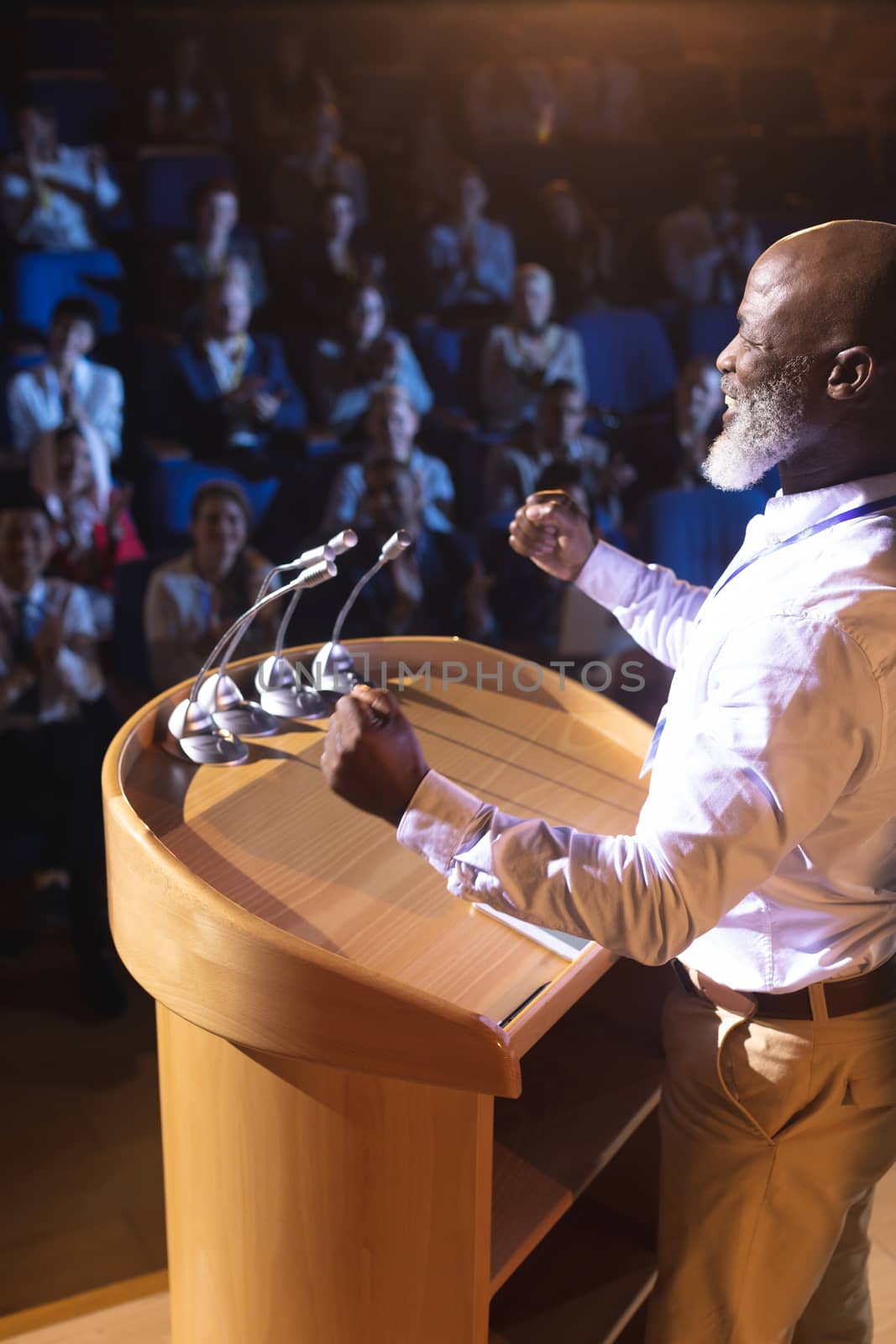 Side view of old African-American businessman standing near podium and giving speech to the audience in the auditorium
