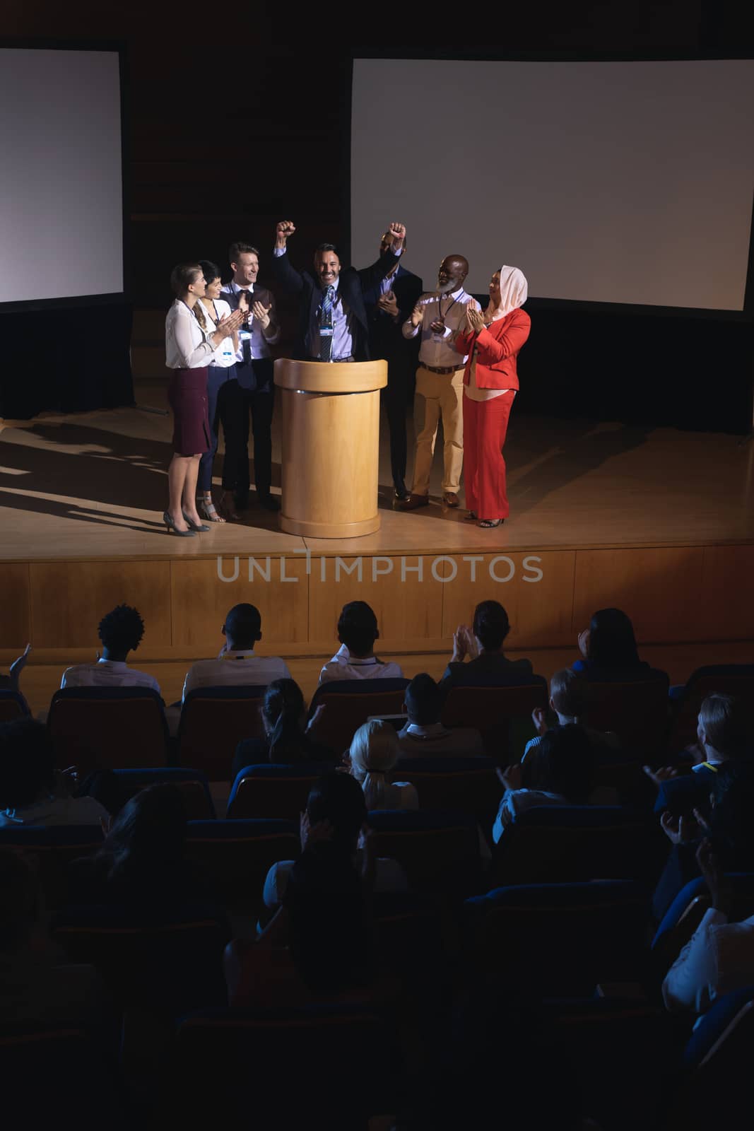 Front view of happy mixed race businessman standing at the stage of the auditorium with colleagues in front of audience