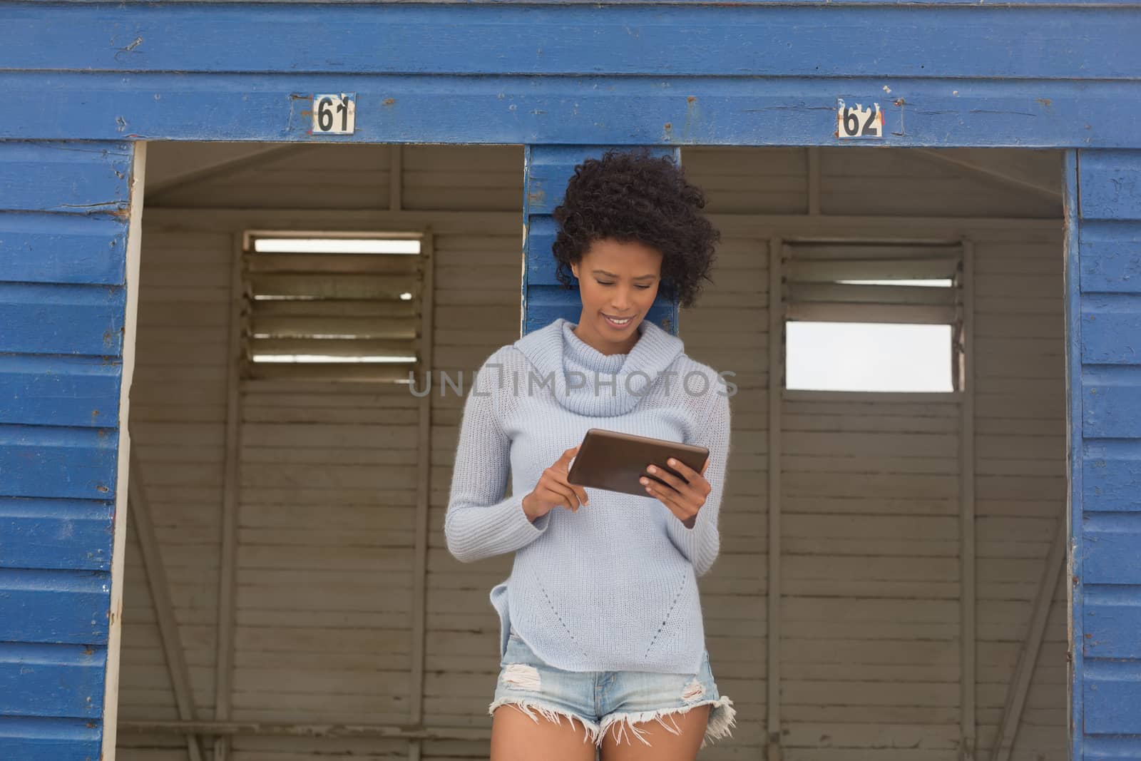 Front view of pretty happy young African American woman using digital tablet standing at beach hut. She is smiling