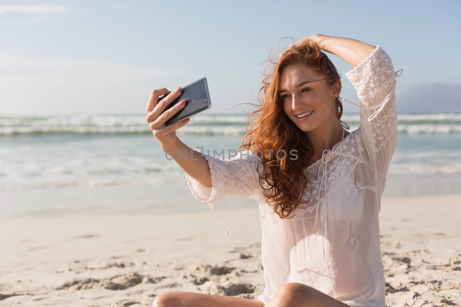 Front view of beautiful happy young woman taking selfie with mobile phone on beach. It is sunny day