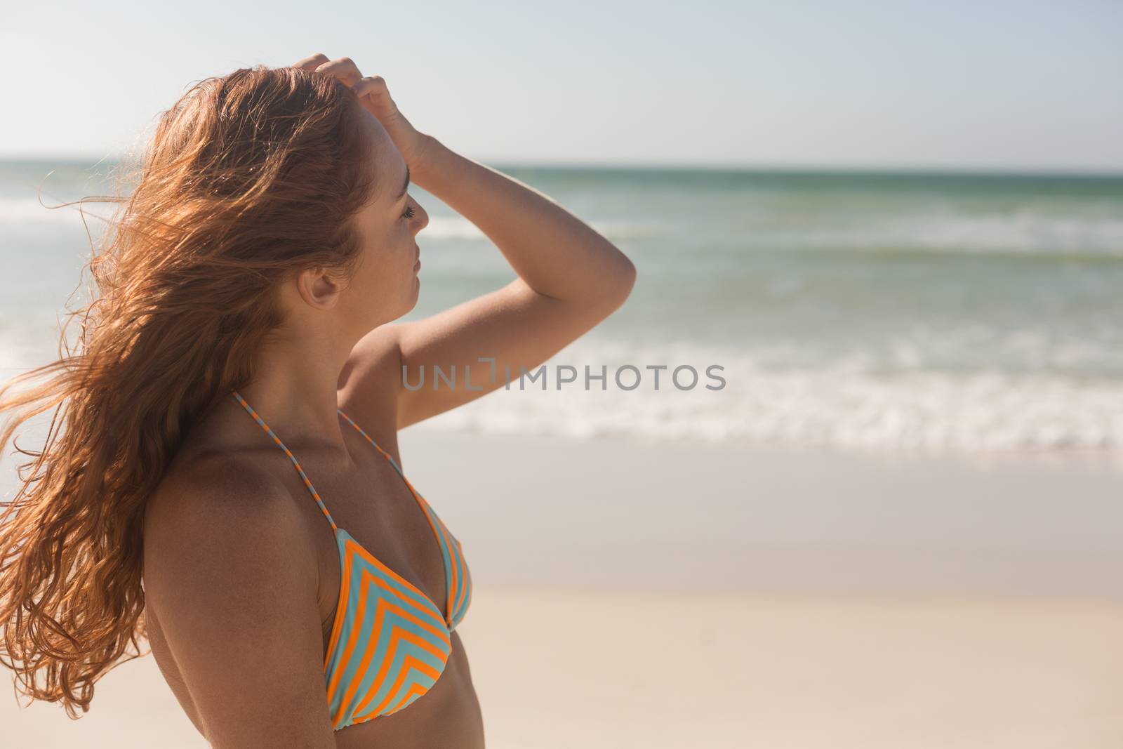 Side view of pretty young Caucasian woman in bikini caressing her hair on the beach. She seems relaxed