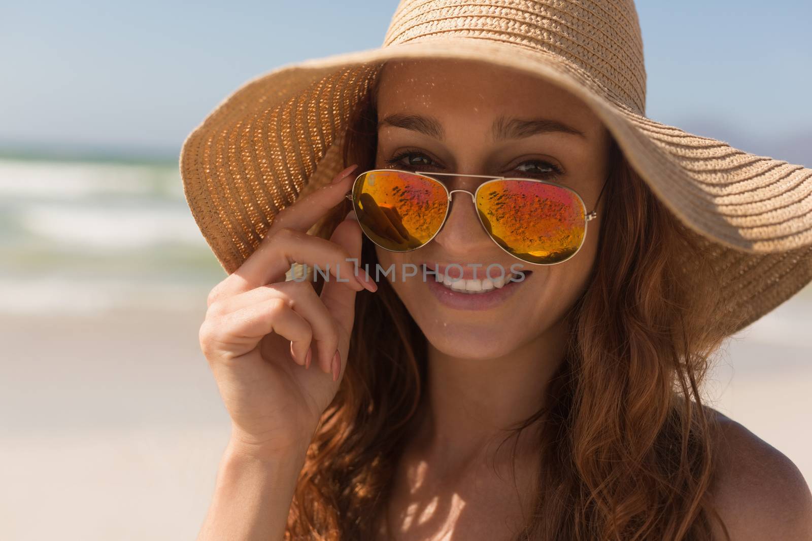 Portrait of pretty young Caucasian woman with hat looking over sunglasses on the beach. She is smiling