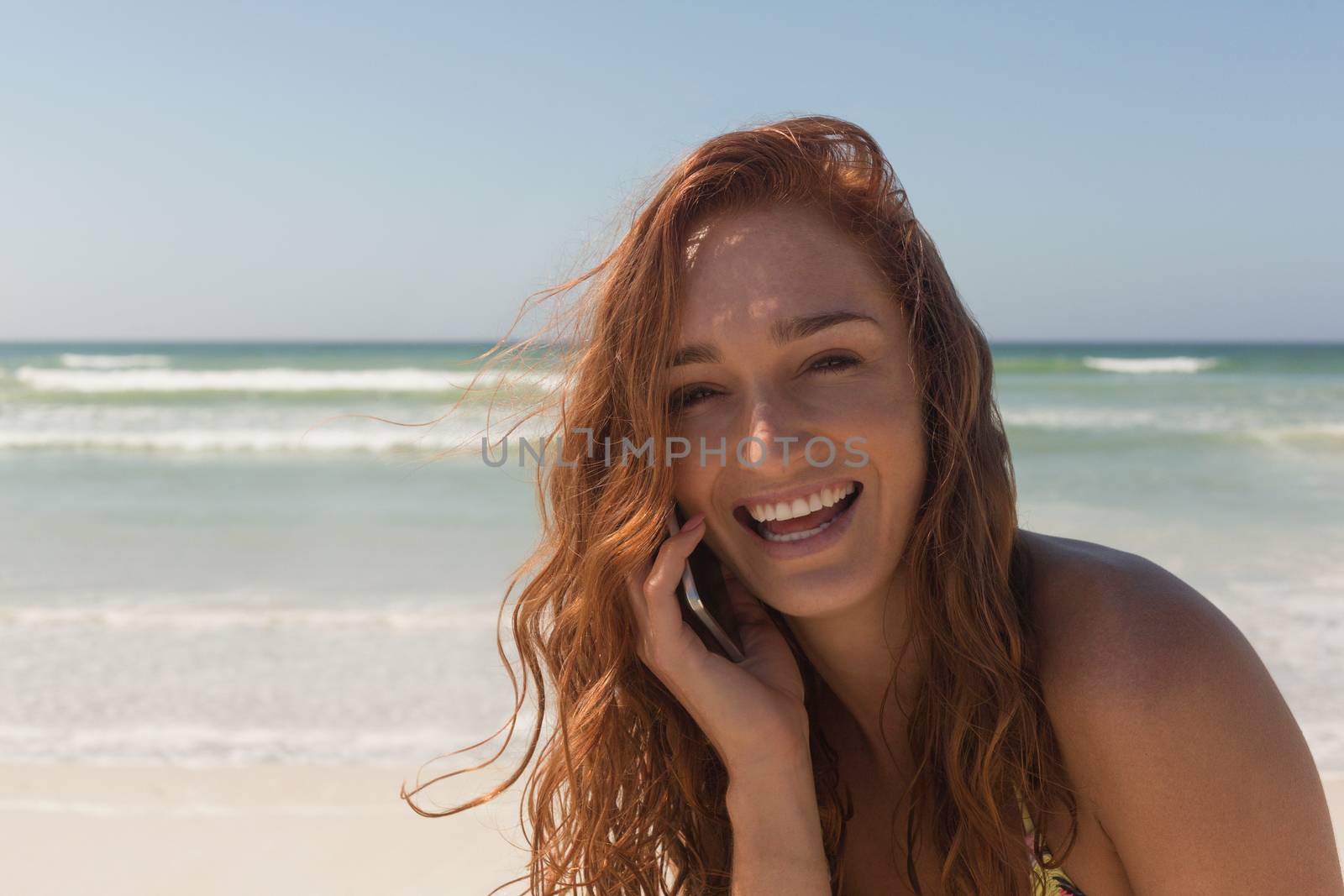 Portrait of happy beautiful young Caucasian woman in bikini talking to her mobile phone at beach. She is smiling and looking at camera