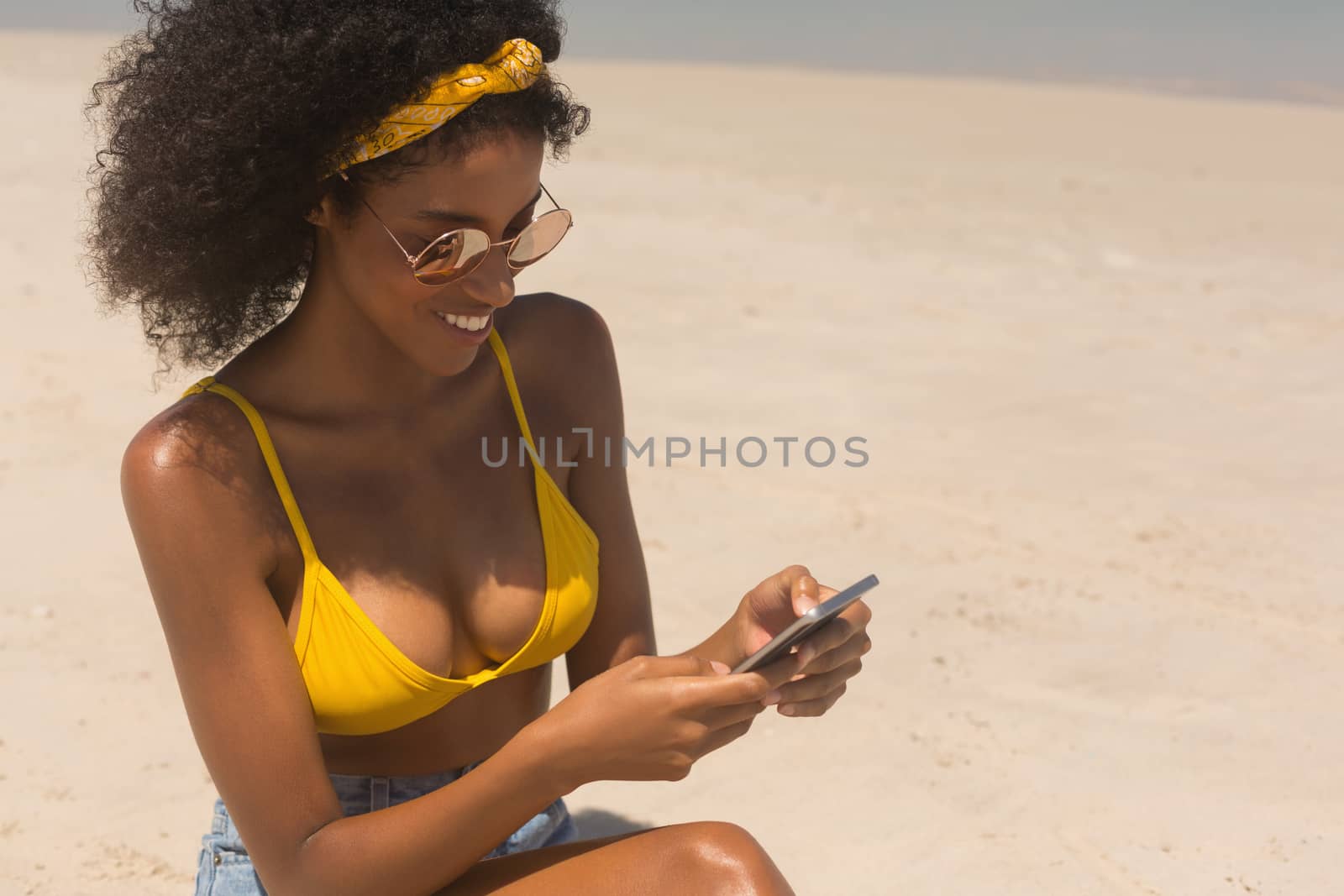 High angle view of pretty young African American woman in yellow bikini and sunglasses using mobile phone sitting on the beach. She is smiling