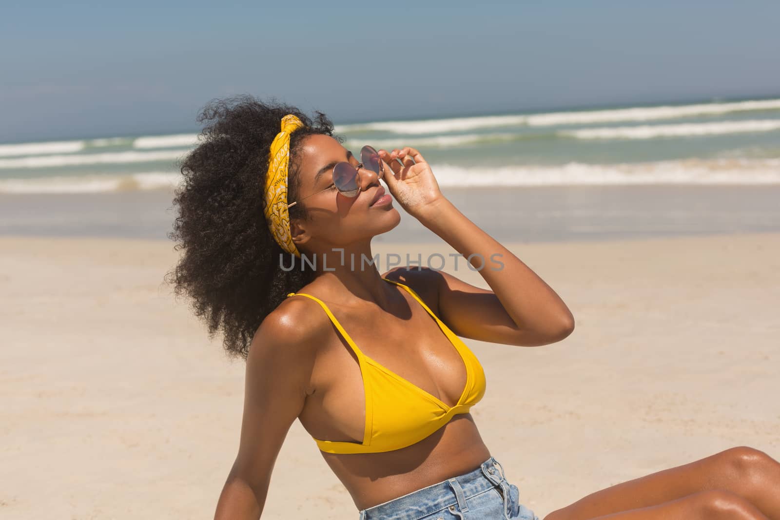 Side view of young African American woman in yellow bikini and sunglasses relaxing on beach in the sunshine. She is enjoying her holidays
