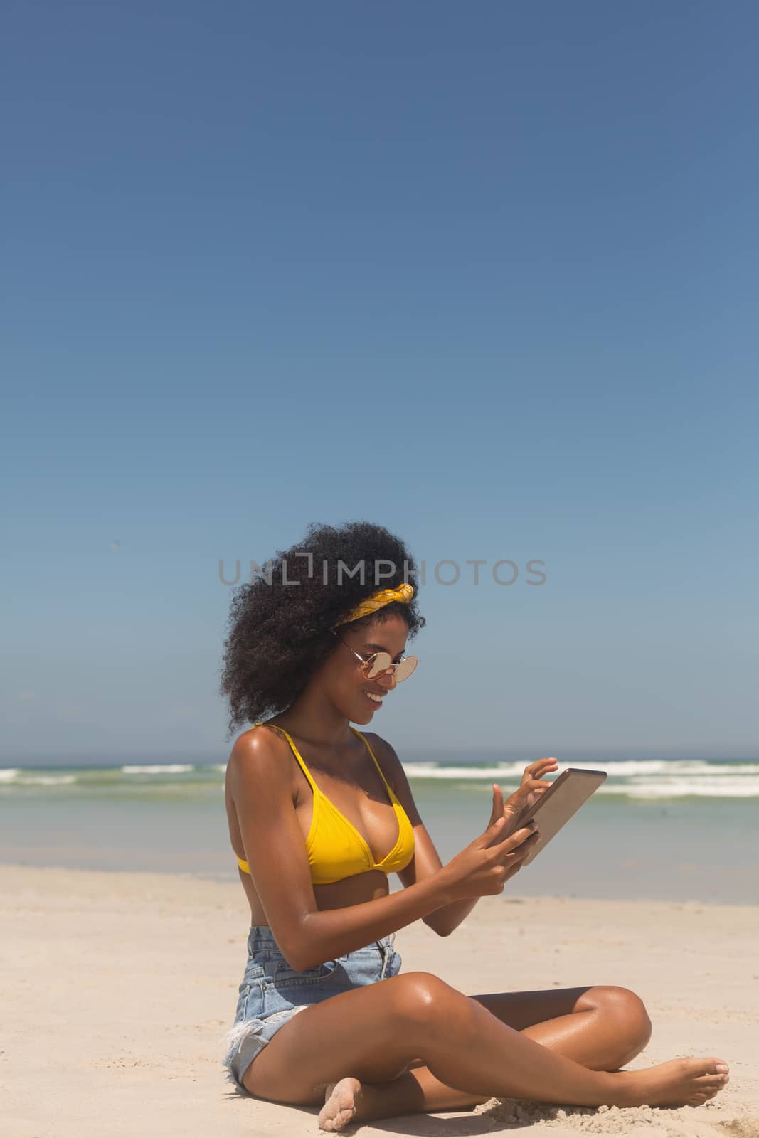 Side view of young African American woman in yellow bikini and sunglasses using digital tablet sitting on the beach. She is smiling