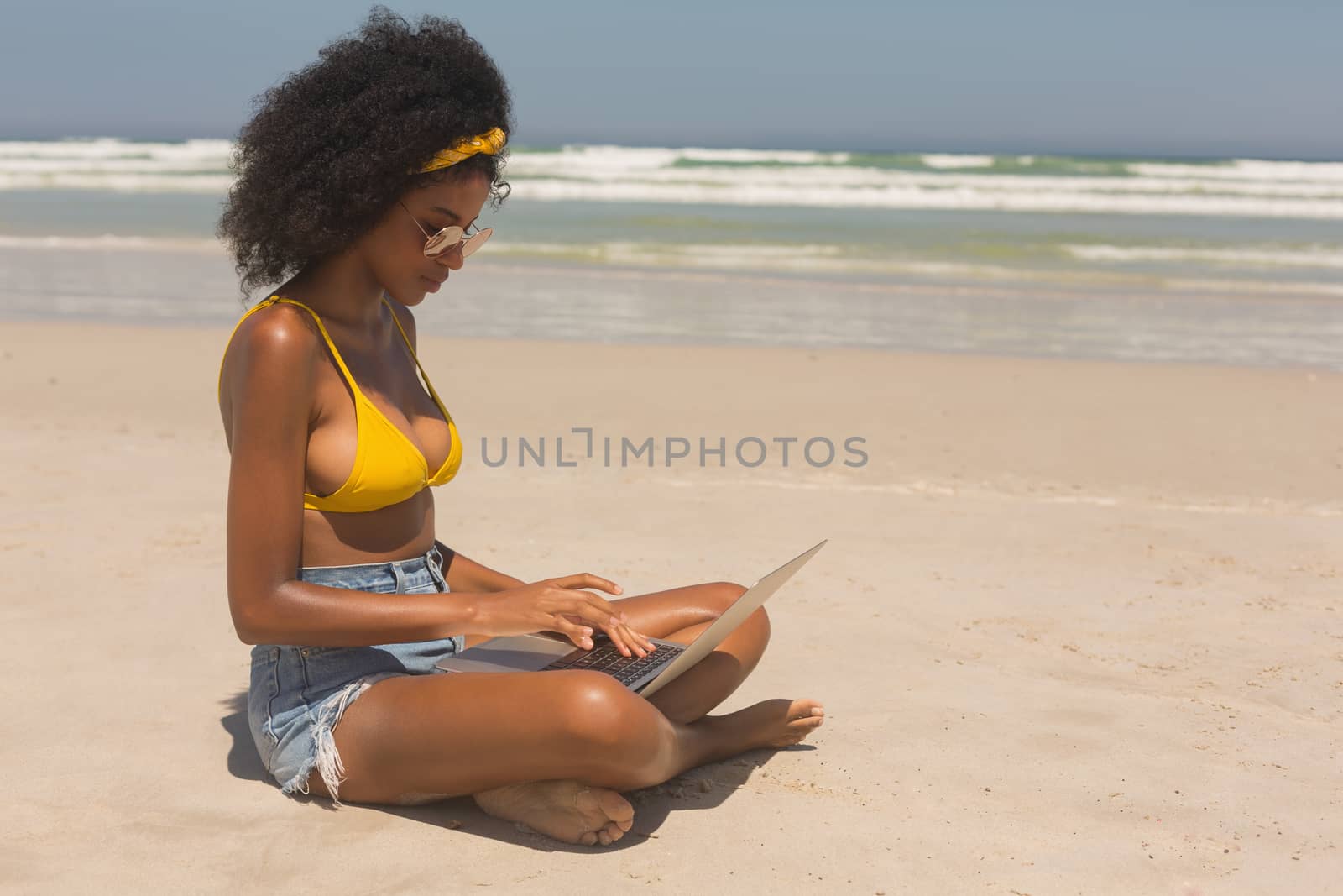 Side view of young African American woman in yellow bikini and sunglasses using laptop sitting on the beach