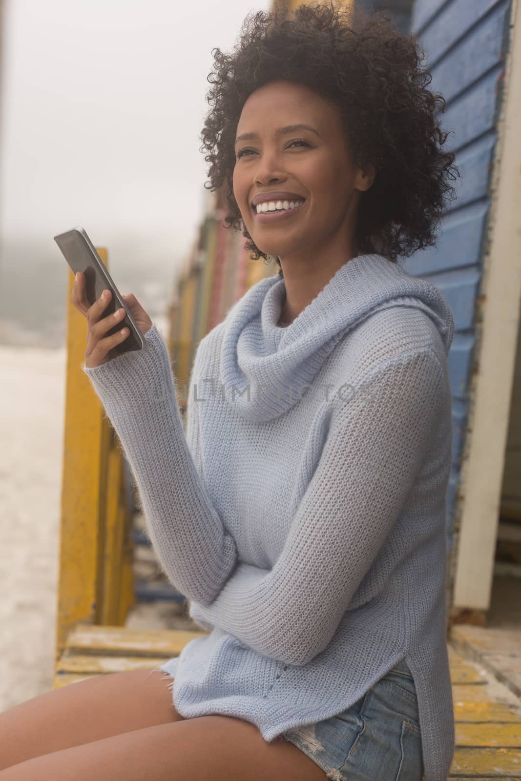 Front view of happy young African American woman using her mobile phone sitting at beach hut. She is smiling