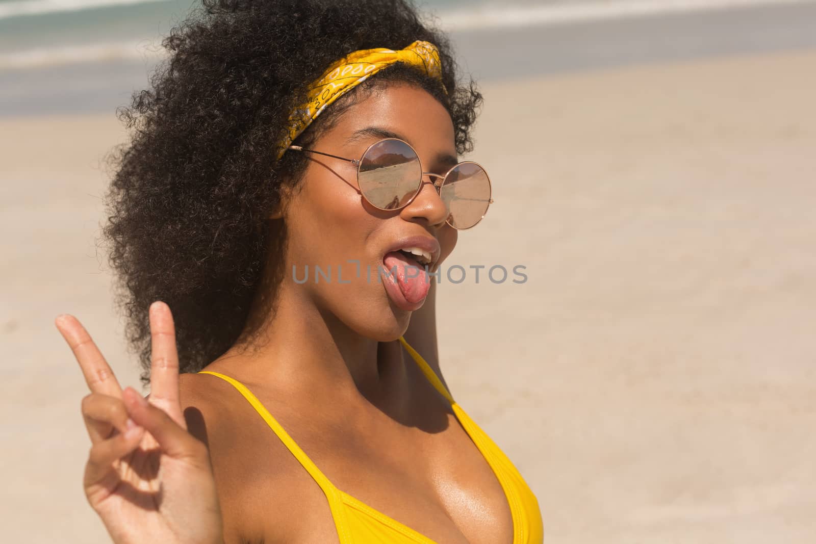 Portrait of young African American woman in bikini and sunglasses shows tongue and victory sign on the beach. She is smiling and looking at camera