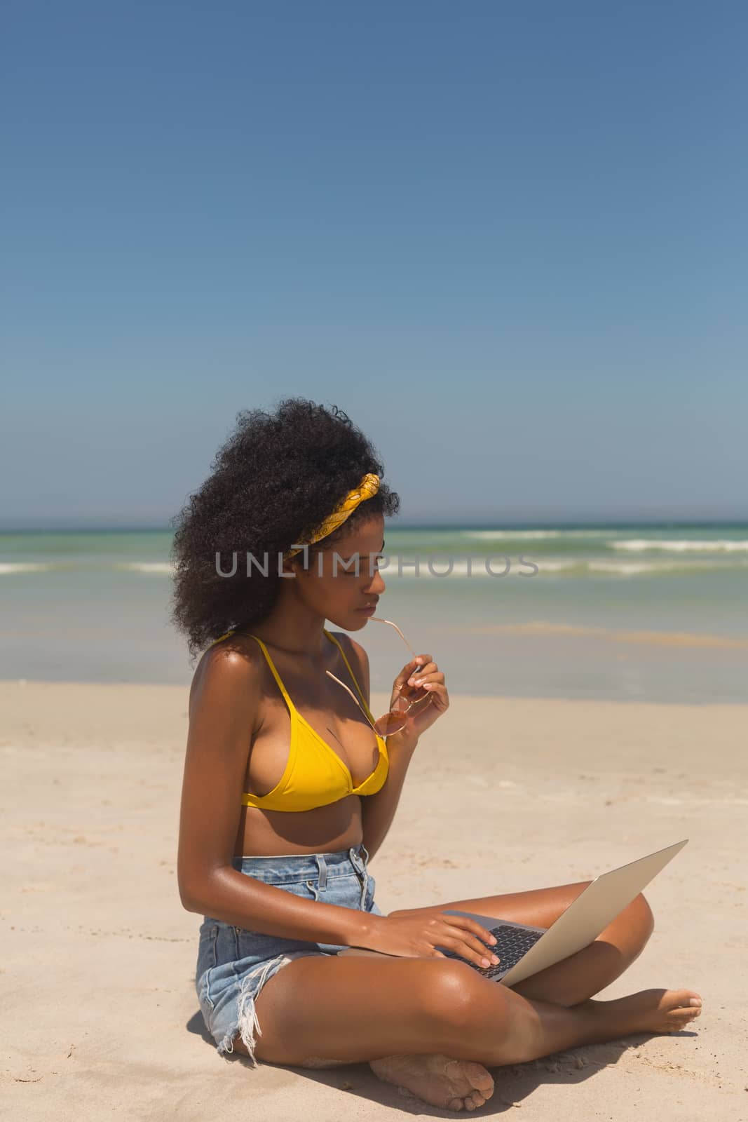 Side view of young African American woman in yellow bikini and sunglasses using laptop sitting on the beach