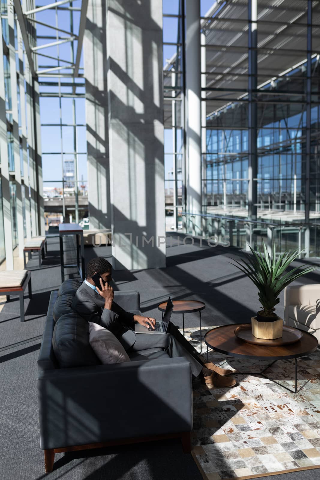 Side view of young African-American businessman talking on mobile phone while using laptop on sofa in modern office