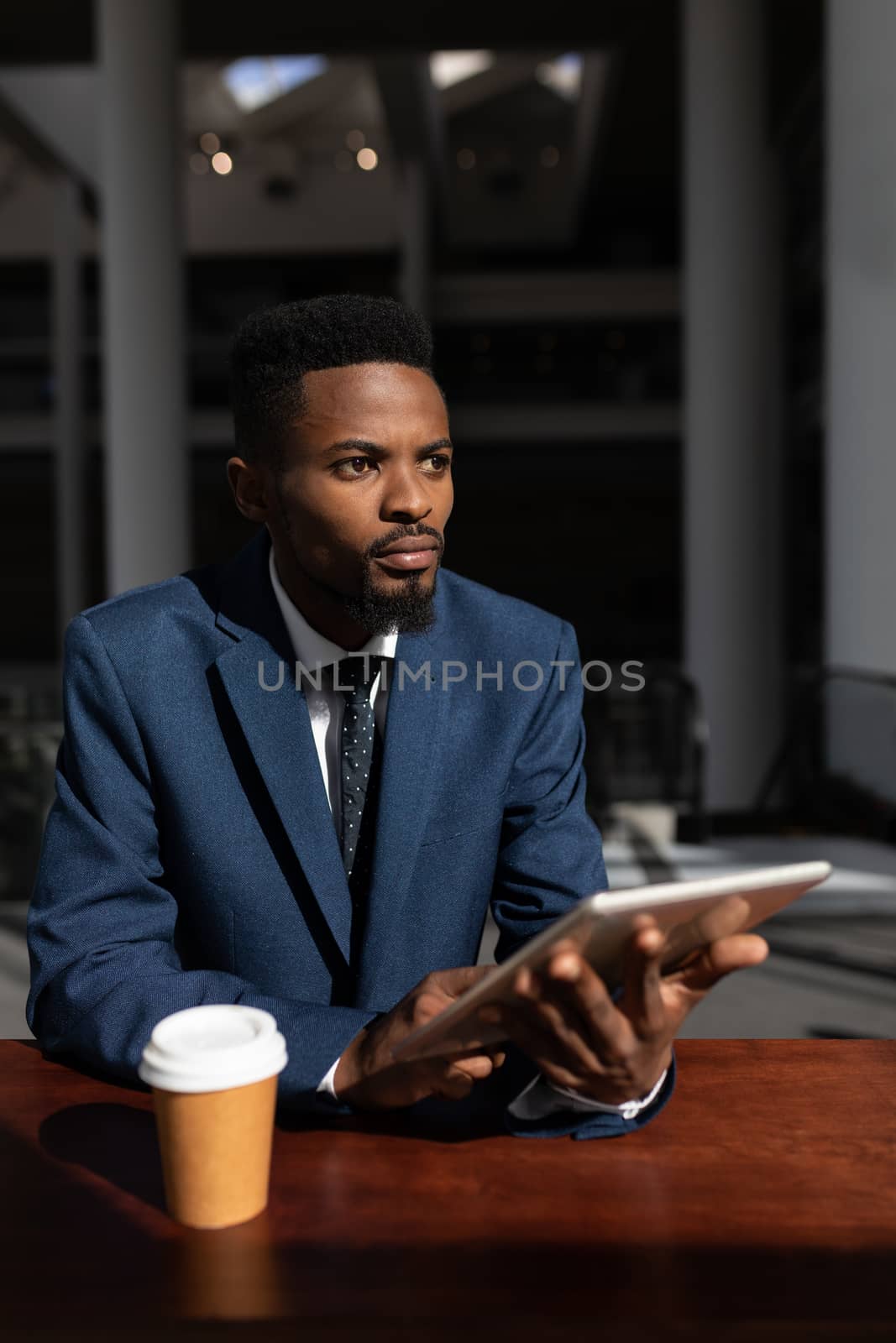 Front view of thoughtful young African-American businessman with digital tablet looking away at table in modern office