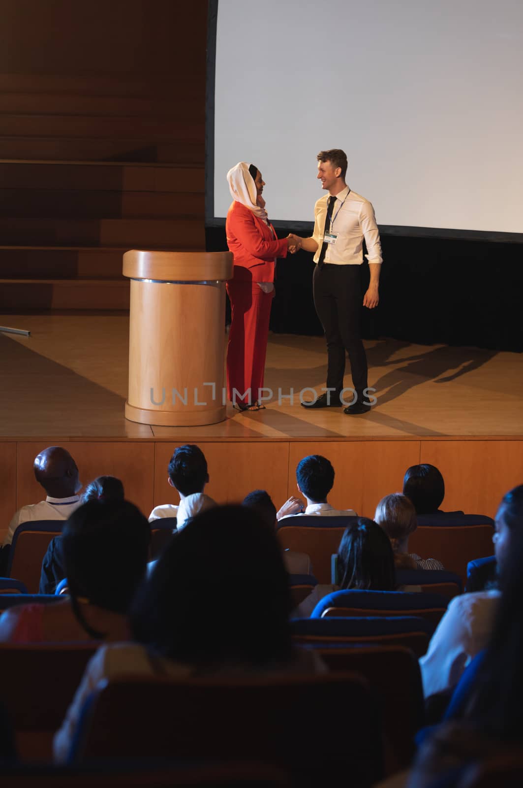 Front view of mixed race business colleague standing and discussing with each other in front of the audience in auditorium while shaking hand 