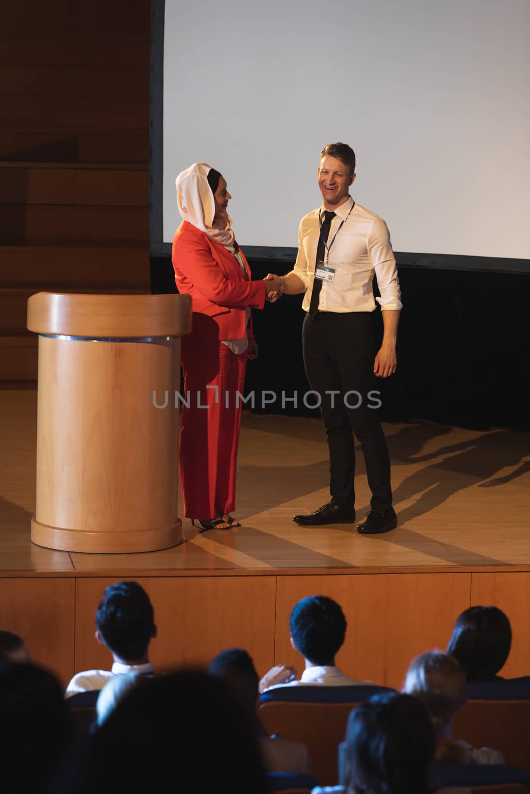 Front view of mixed race business colleague standing and discussing with each other in front of the audience in auditorium while shaking hand 