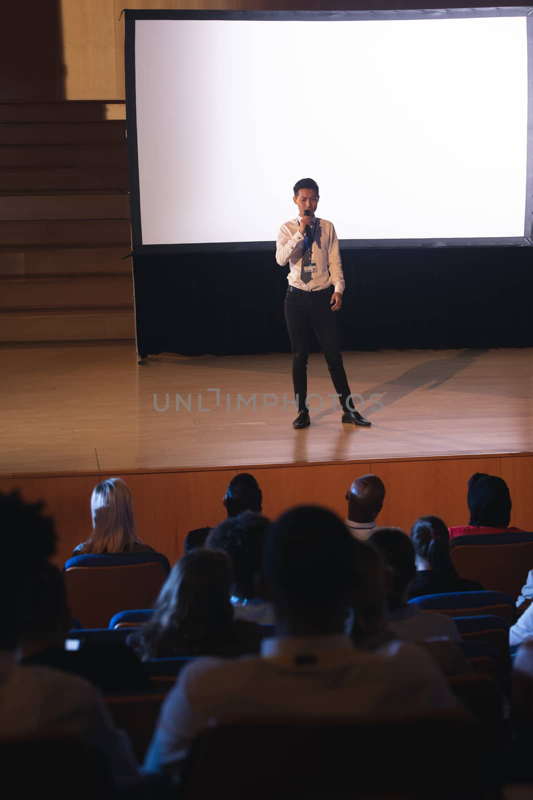 High view of young Asian businessman standing and giving presentation in auditorium while holding mike in his hand in auditorium