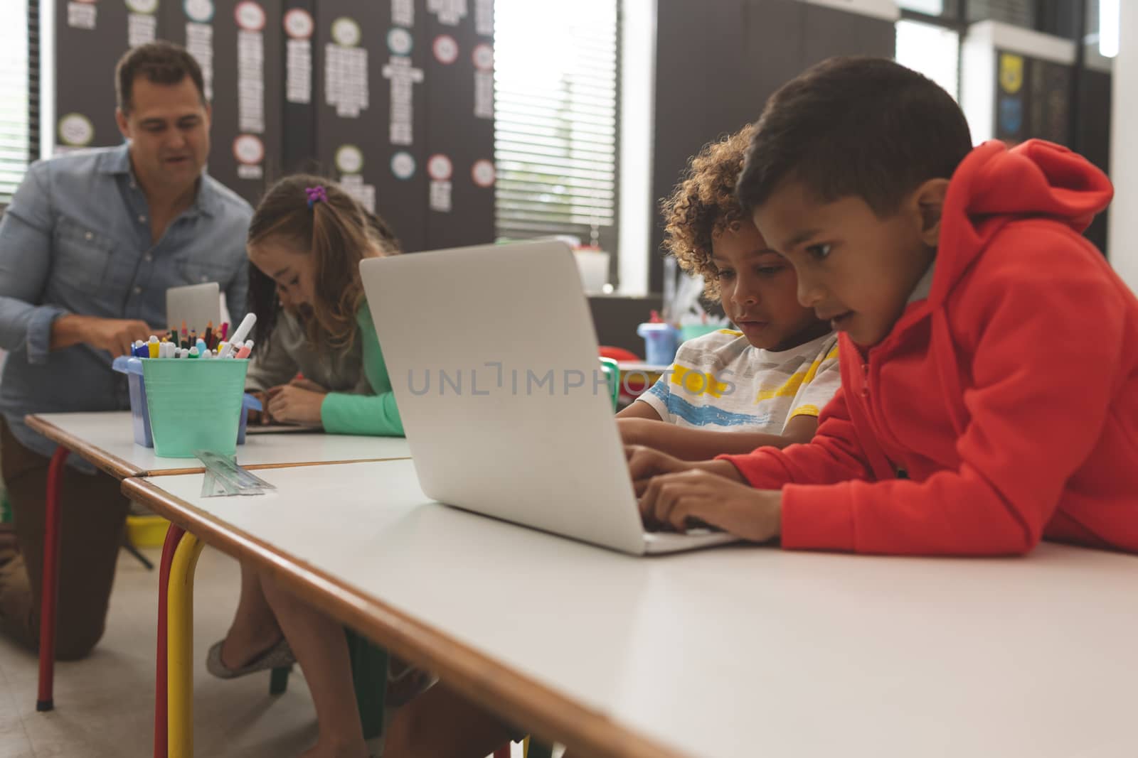 Side view of two mixed-race school boys working and scrolling on one laptop while their teacher teaching to another school girl in background