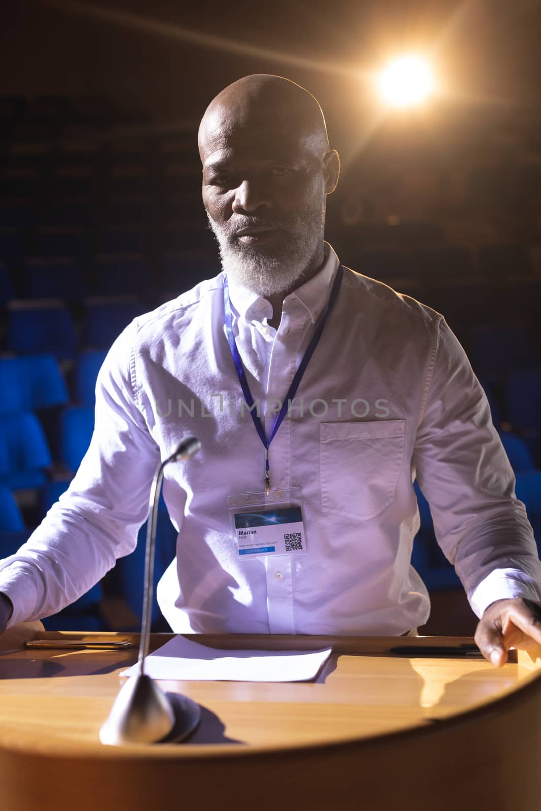 Front view of African-American usinessman standing at podium on stage in auditorium  while looking at camera with orange light in hte background